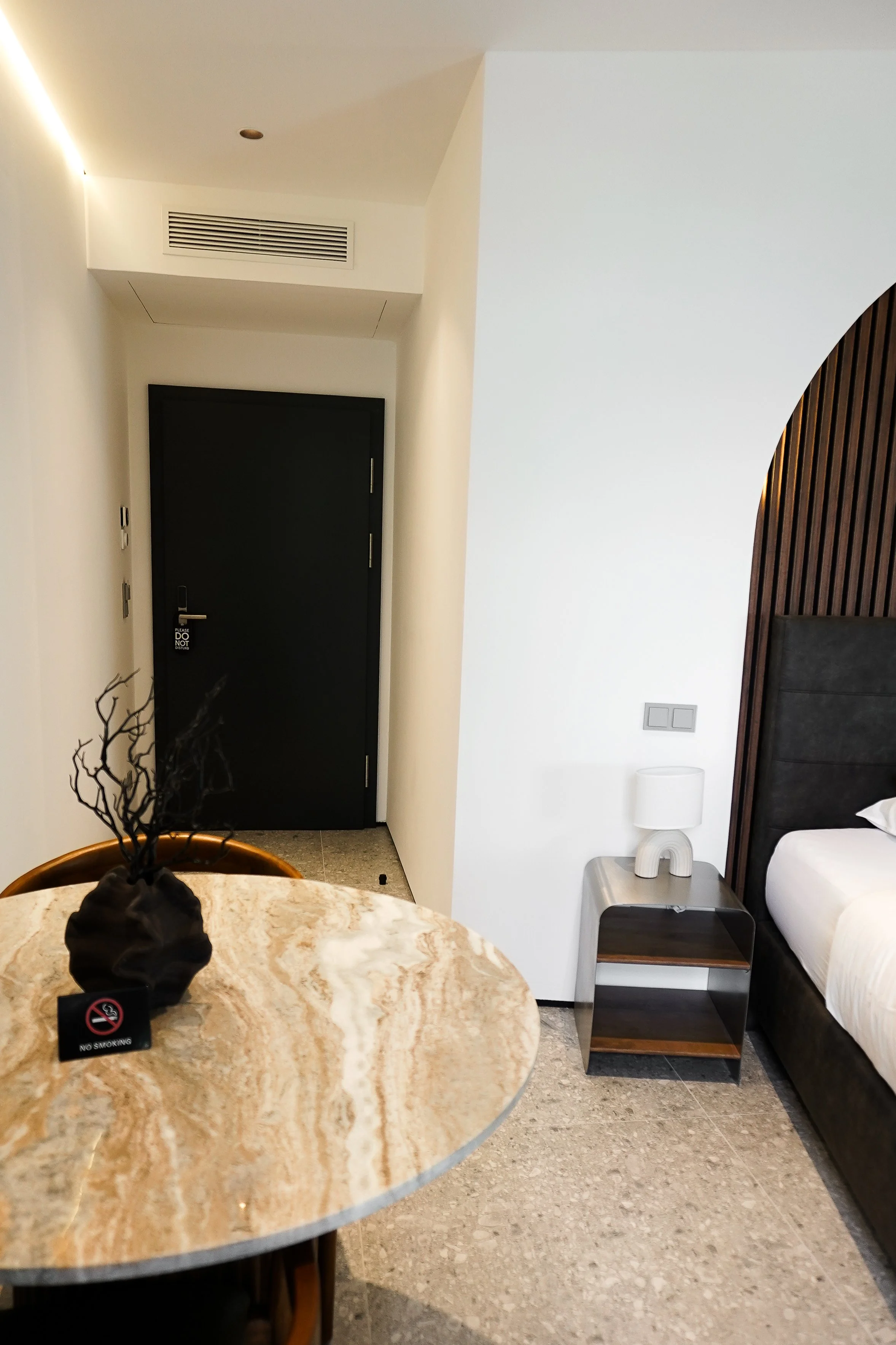 Modernes Hotelzimmer mit schwarzer Tür, Nachttisch, Lampe, Bett, runder Marmortisch mit Dekorationsvase und 'No Smoking'-Schild.