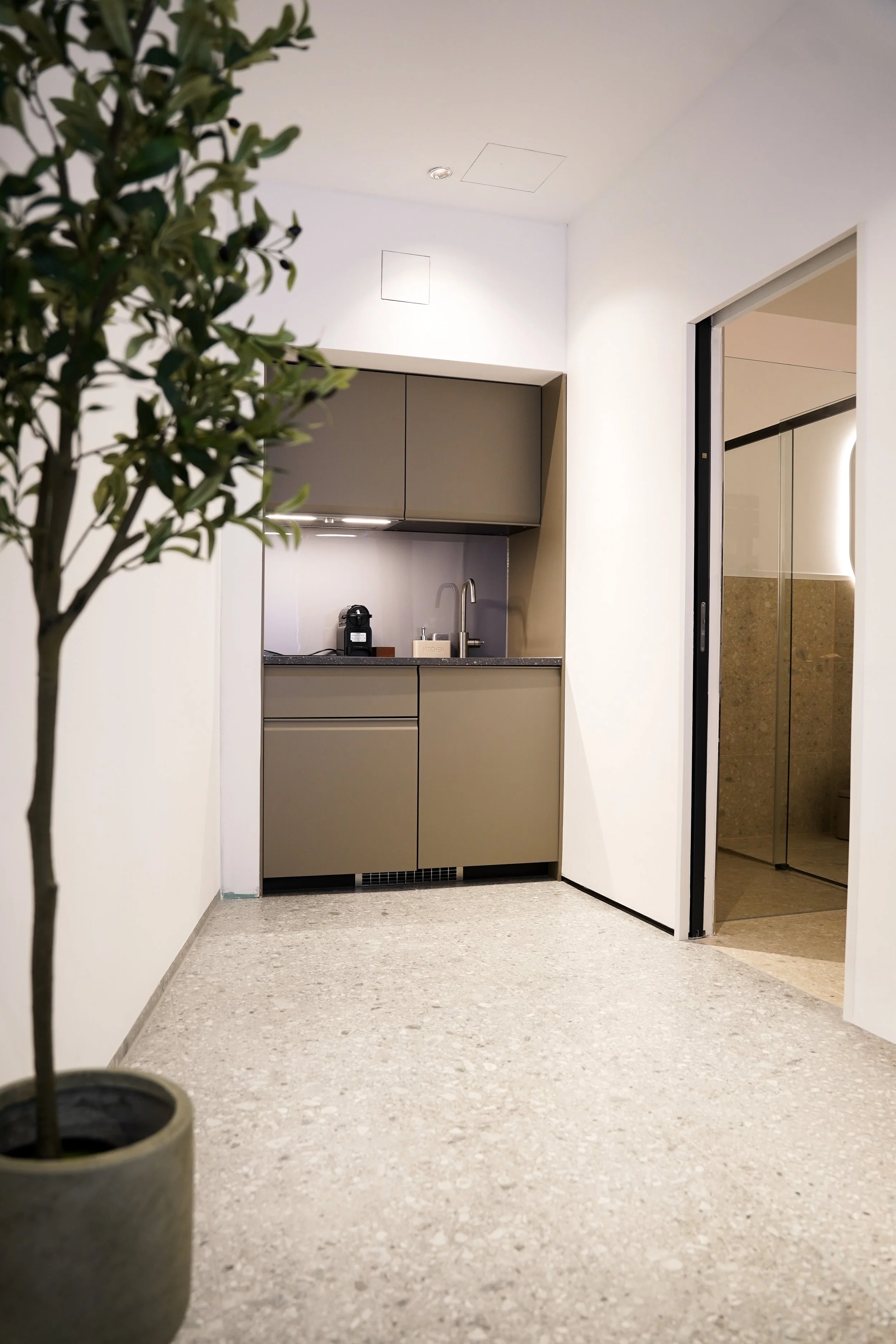 Kleine Küche mit grauen Schränken, Wasserkocher, Kaffeemaschine und Spüle, daneben ein offener Raum mit Glastür