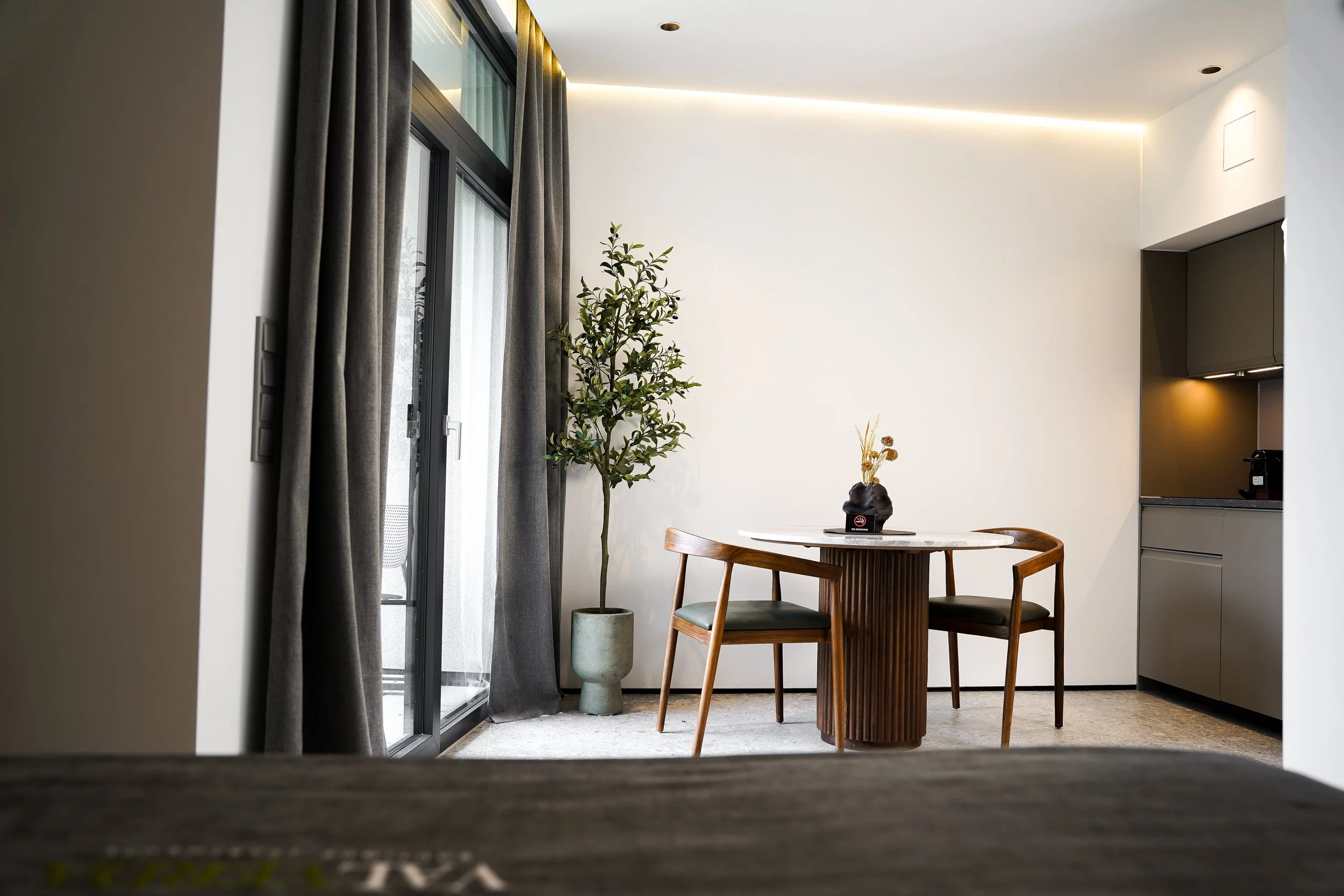 Ein Wohnzimmer mit einem runden Holztisch, zwei Stühlen, einer Topfpflanze neben einem Fenster mit dunklen Vorhängen, eine moderne Küchenzeile im Hintergrund, hell erleuchtet mit indirekter Beleuchtung.