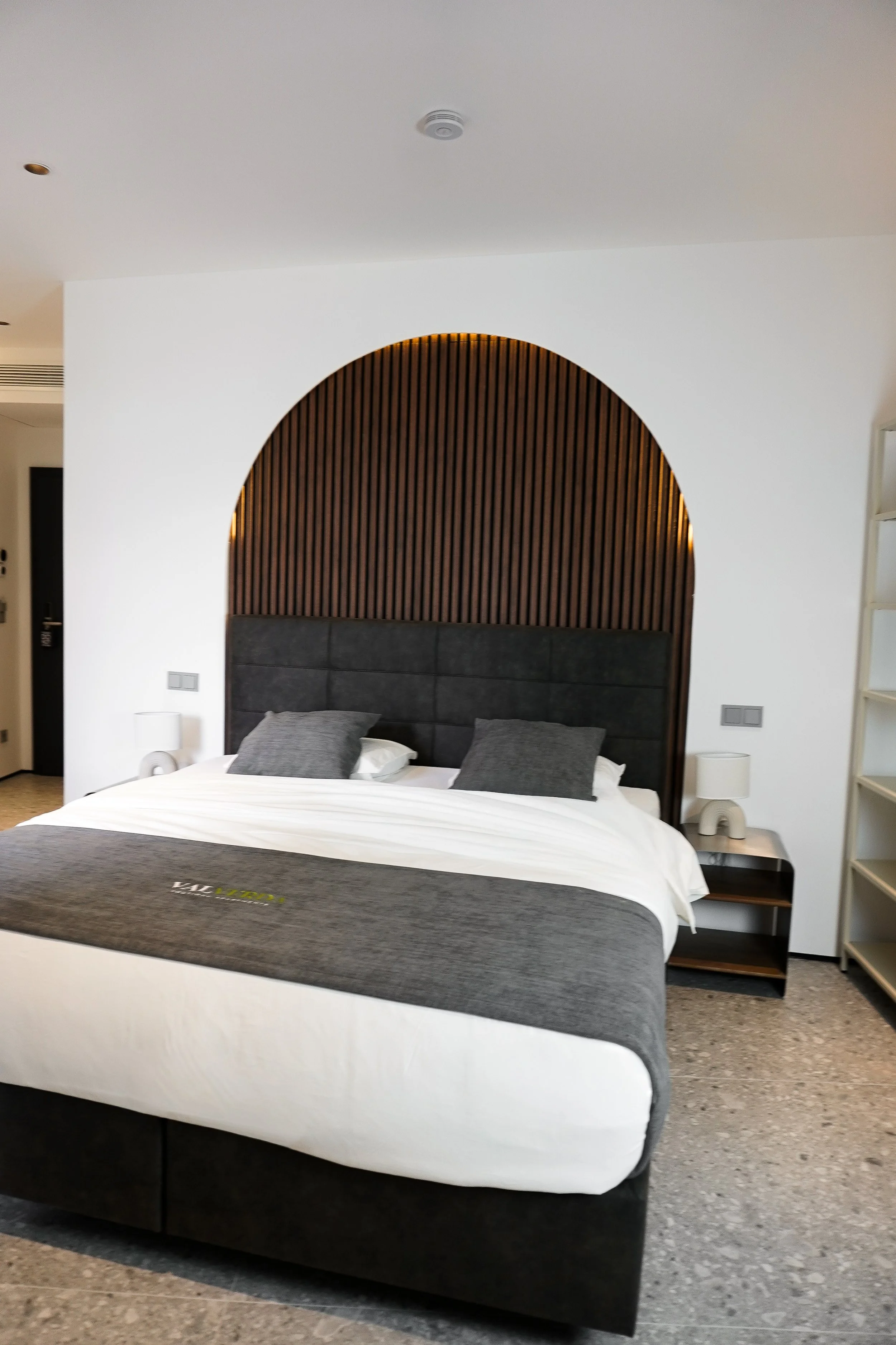 Ein modernes Hotelzimmer mit einem Bett, schwarzer Kopfleiste, grauen Kissen, weißen Bettwäsche, seitlichen Nachttischen und Nachttischlampen, im Hintergrund eine Wand mit einem halbrunden Holzdesign.