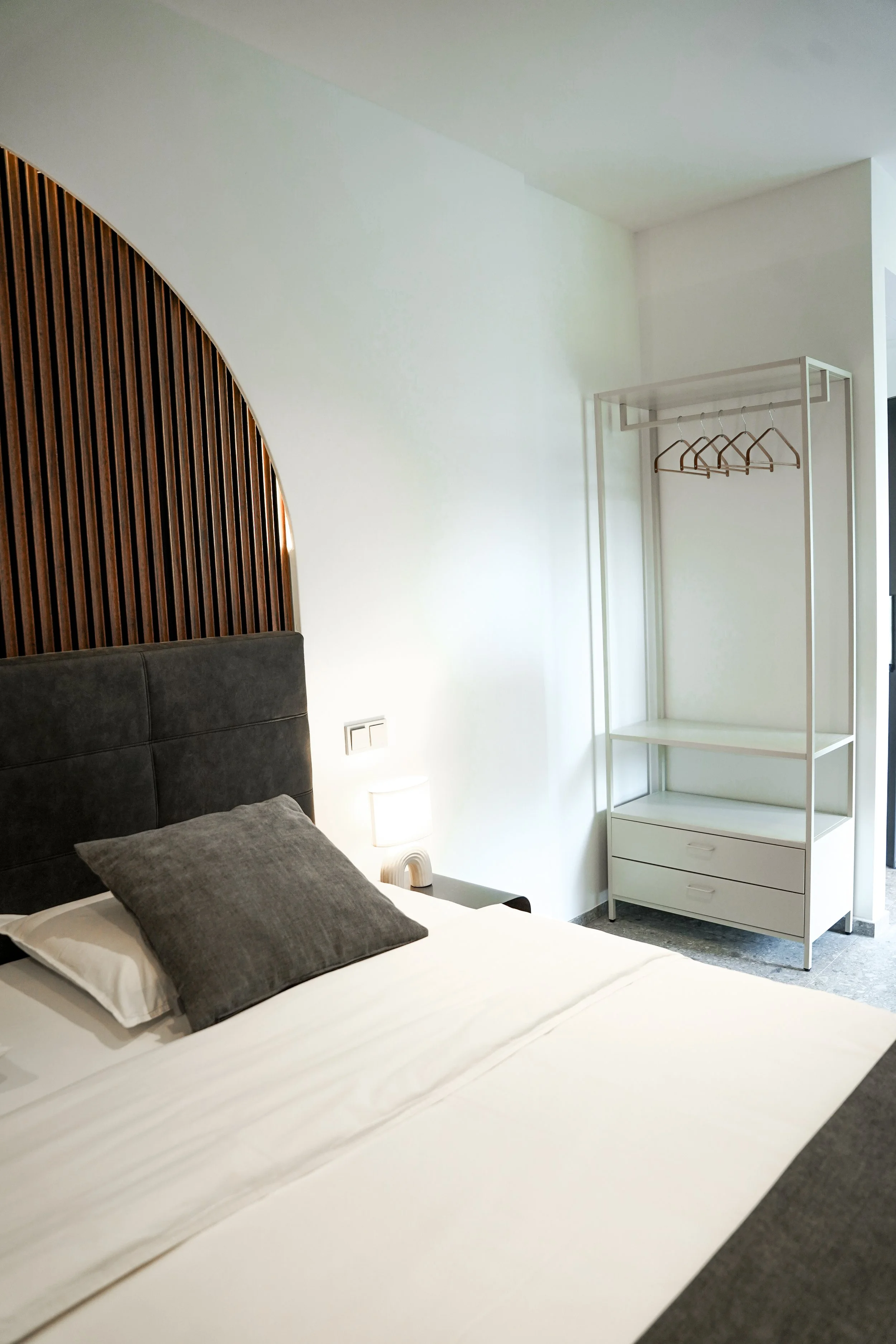 Ein Schlafzimmer mit einem Bett, einem schwarzen gepolsterten Kopfteil, grauem Kissen, Nachttisch, Lampe, und einem weißen offenen Kleiderständer mit Kleiderbügel.