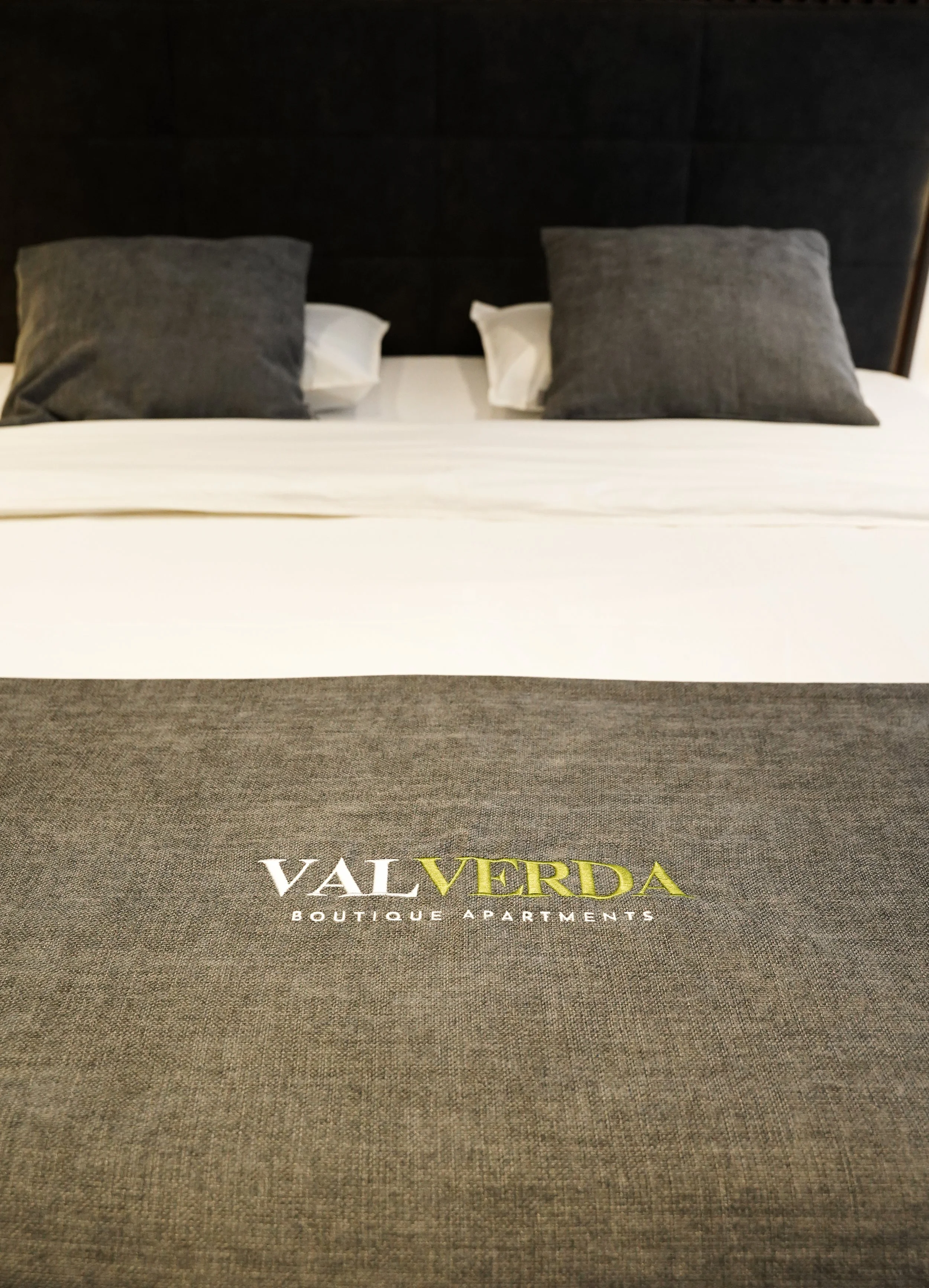 Bett mit weißen Laken, zwei graue Kissen, zwei weiße Kissen, auf der Bettdecke steht 'Valverda Boutique Apartments'.