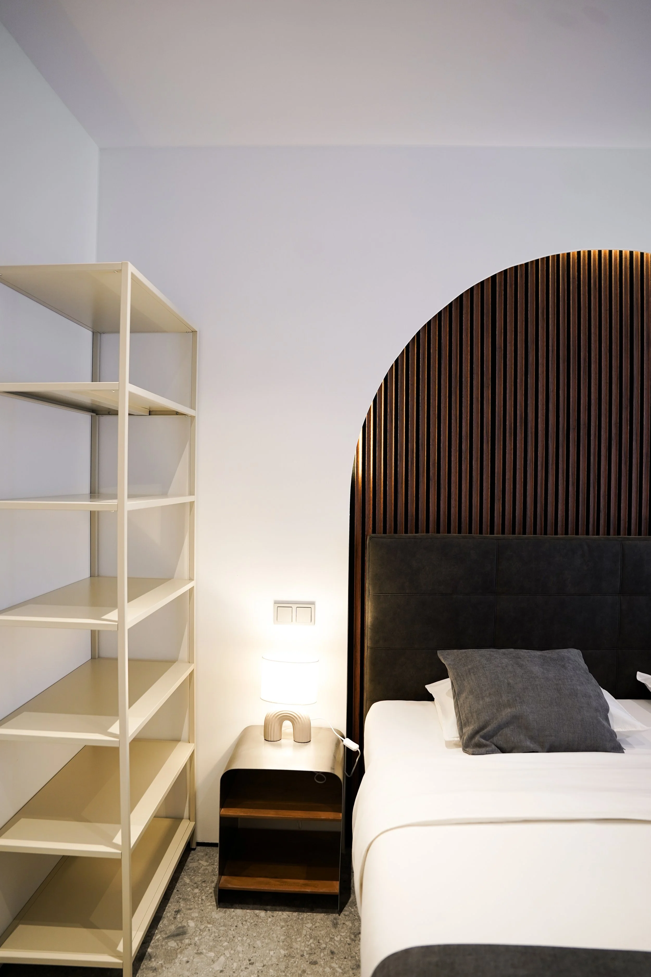 Modernes Schlafzimmer mit schwarzem Kopfteil, grauem Kissen, Nachttisch mit Lampe, leerem weißen Regal, dunklem Laminatboden und weißem Wandhintergrund.