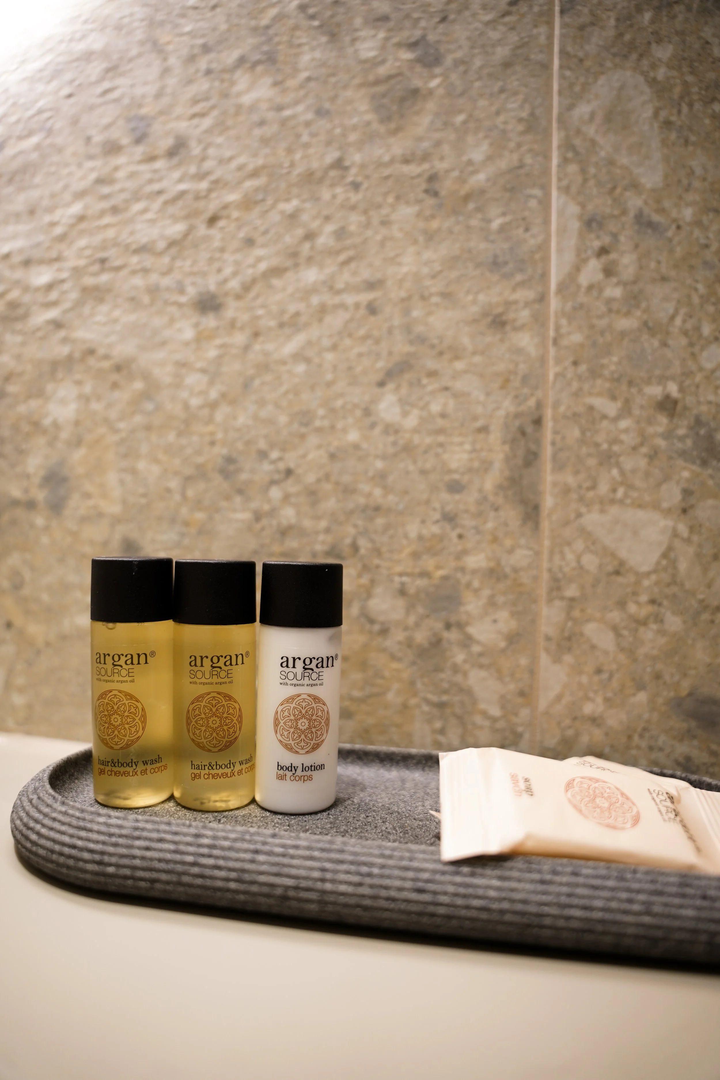 Reise- oder Hotelzimmer mit drei Argan-Produkten auf einer grauen Verpackung, gegen eine steinartige Wand. Zwei kleine Gel-Flaschen und eine Body-Lotion. Paket liegt neben den Flaschen.