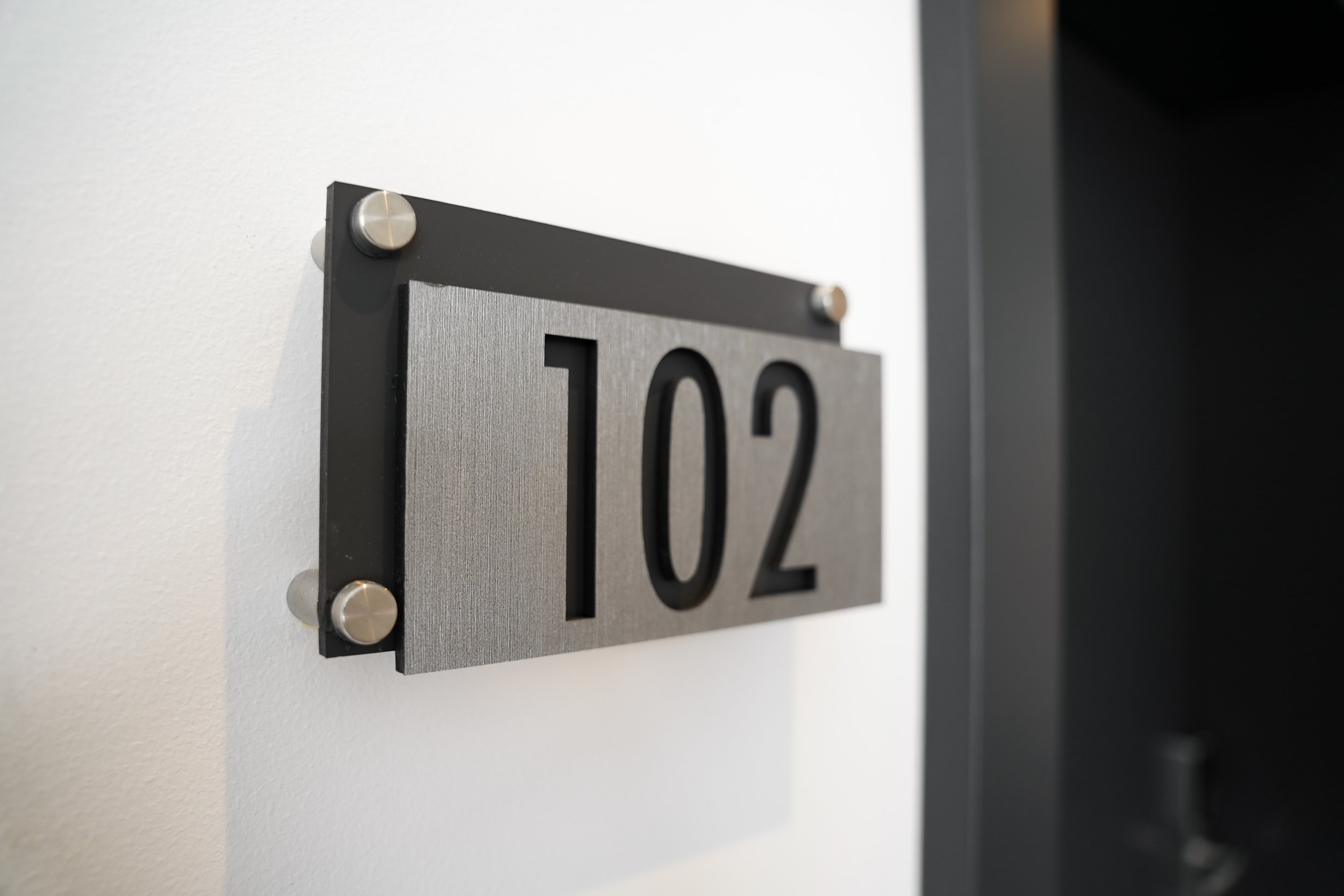 Moderne Zimmernummer 102 an einer weißen Wand, aus Metall mit schwarzen Zahlen.