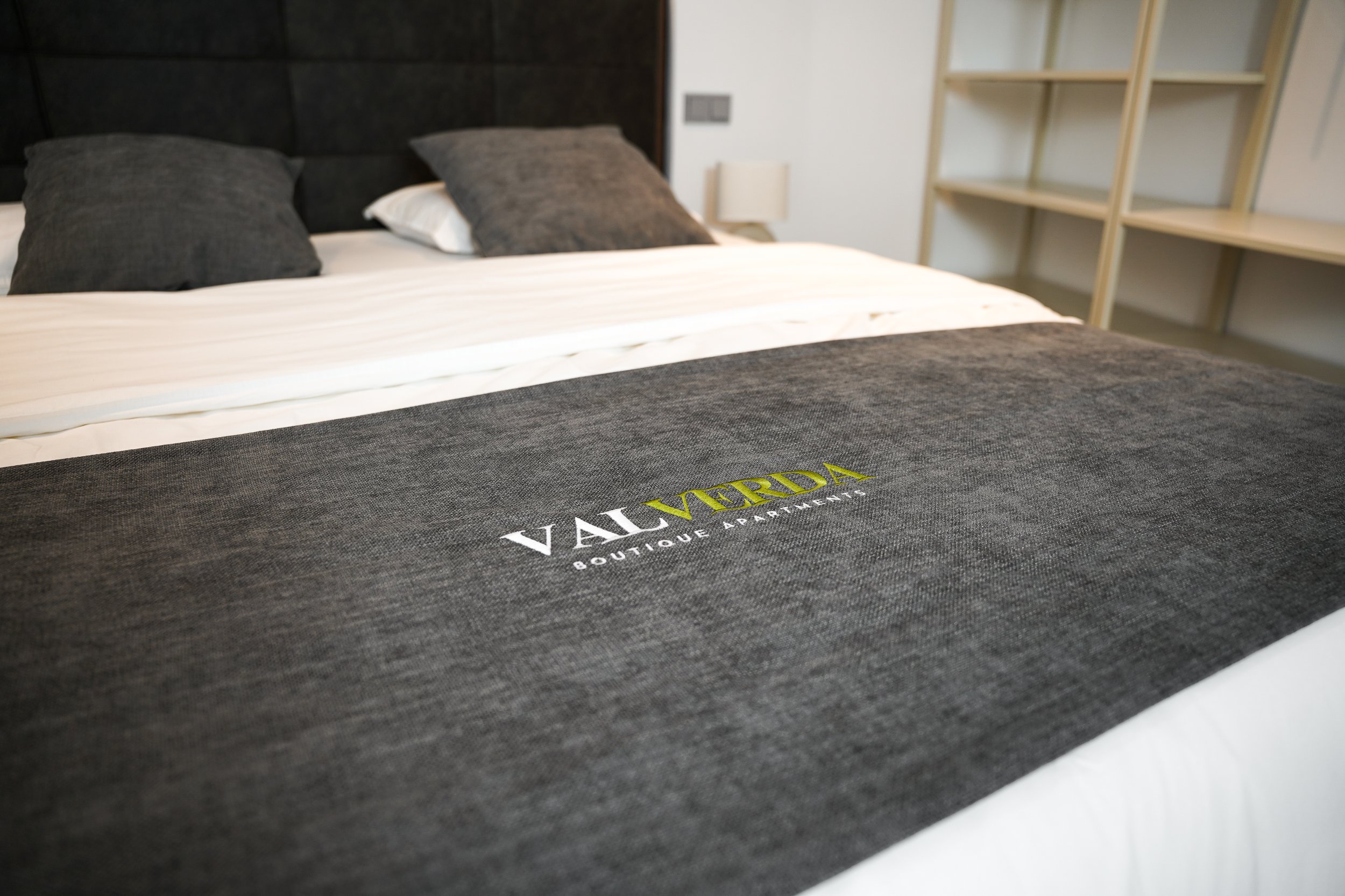 Bett mit schwarzen Kissen, grauer Bettdecke mit VAI MERDA Boutique Apartments Logo, Part of a modern Schlafzimmer eingerichtet mit Regal und Nachttisch.