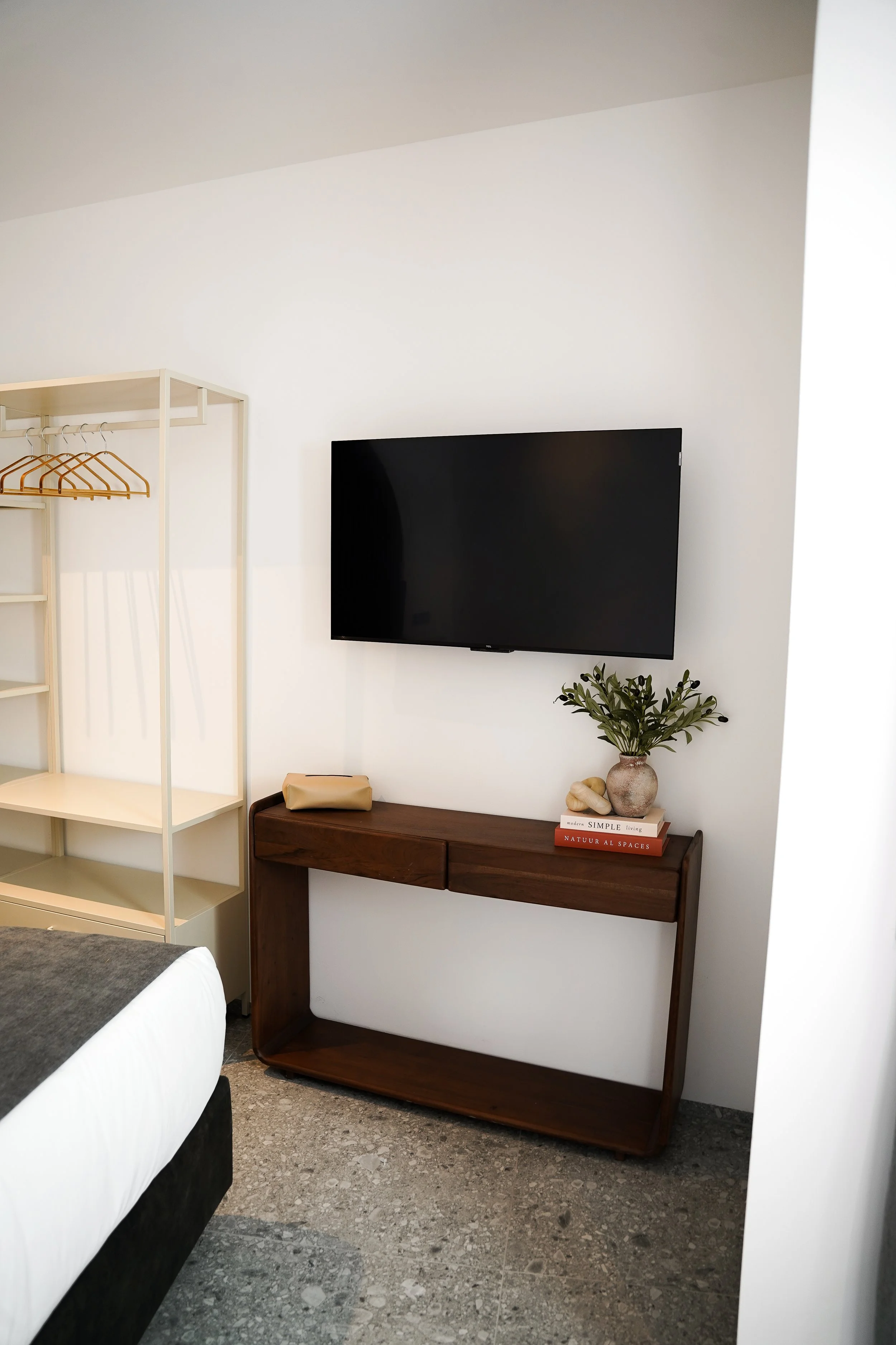 Moderner Wohnraum mit Wandfernseher, dunklem Holzkonsole, grüner Pflanze in Vase, Büchern und Dekoration auf der Konsole, offenem Kleiderschrank mit Kleiderbügel, Teil eines Bettes im Vordergrund, grauem Boden.