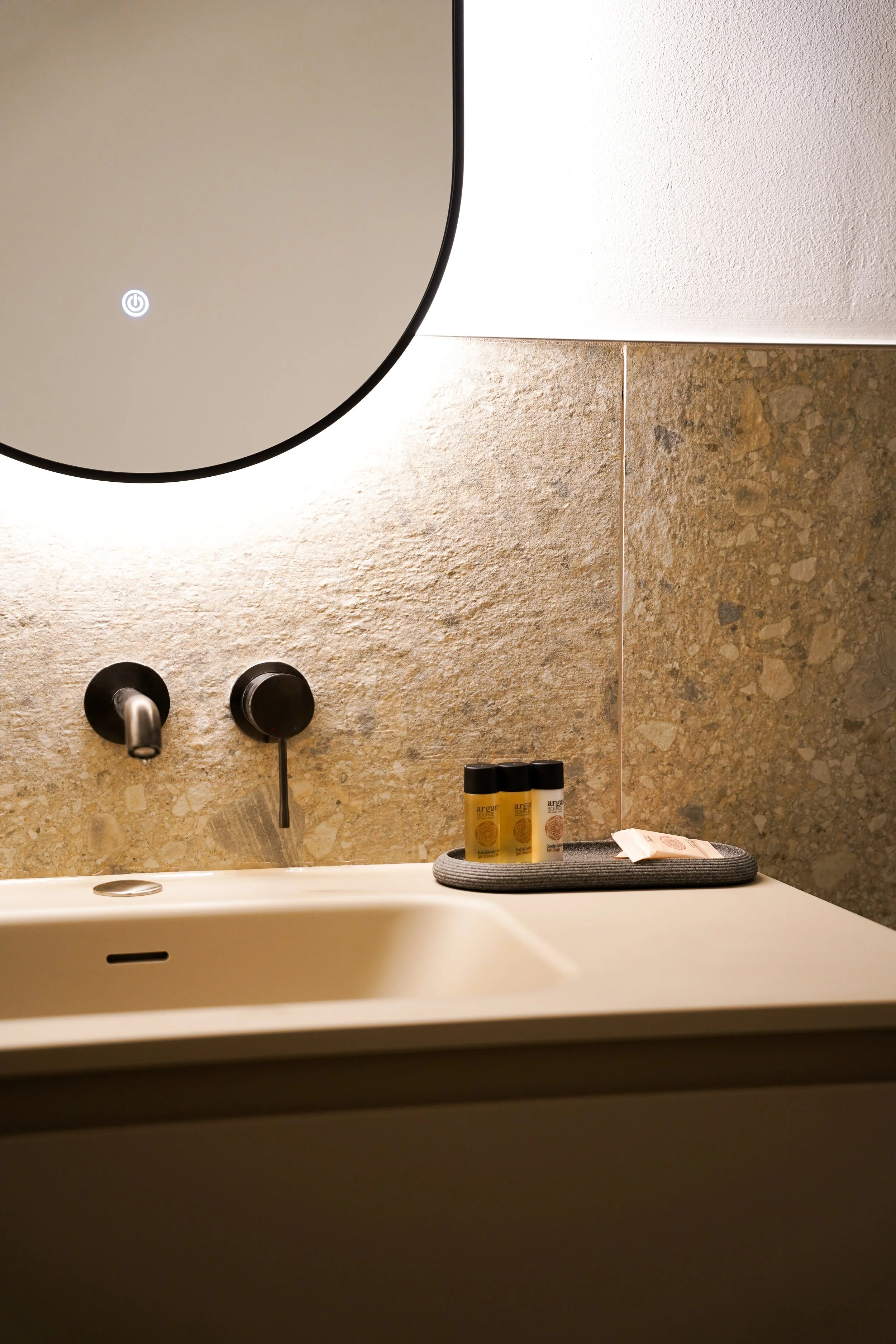 Eine moderne Badezimmerwaschbecken mit Spiegel, schwarzen Wasserhähnen, grauem Handtuch und kleineren Pflegeprodukten auf dem Waschtisch, mit einer sandfarbenen Wand im Hintergrund.