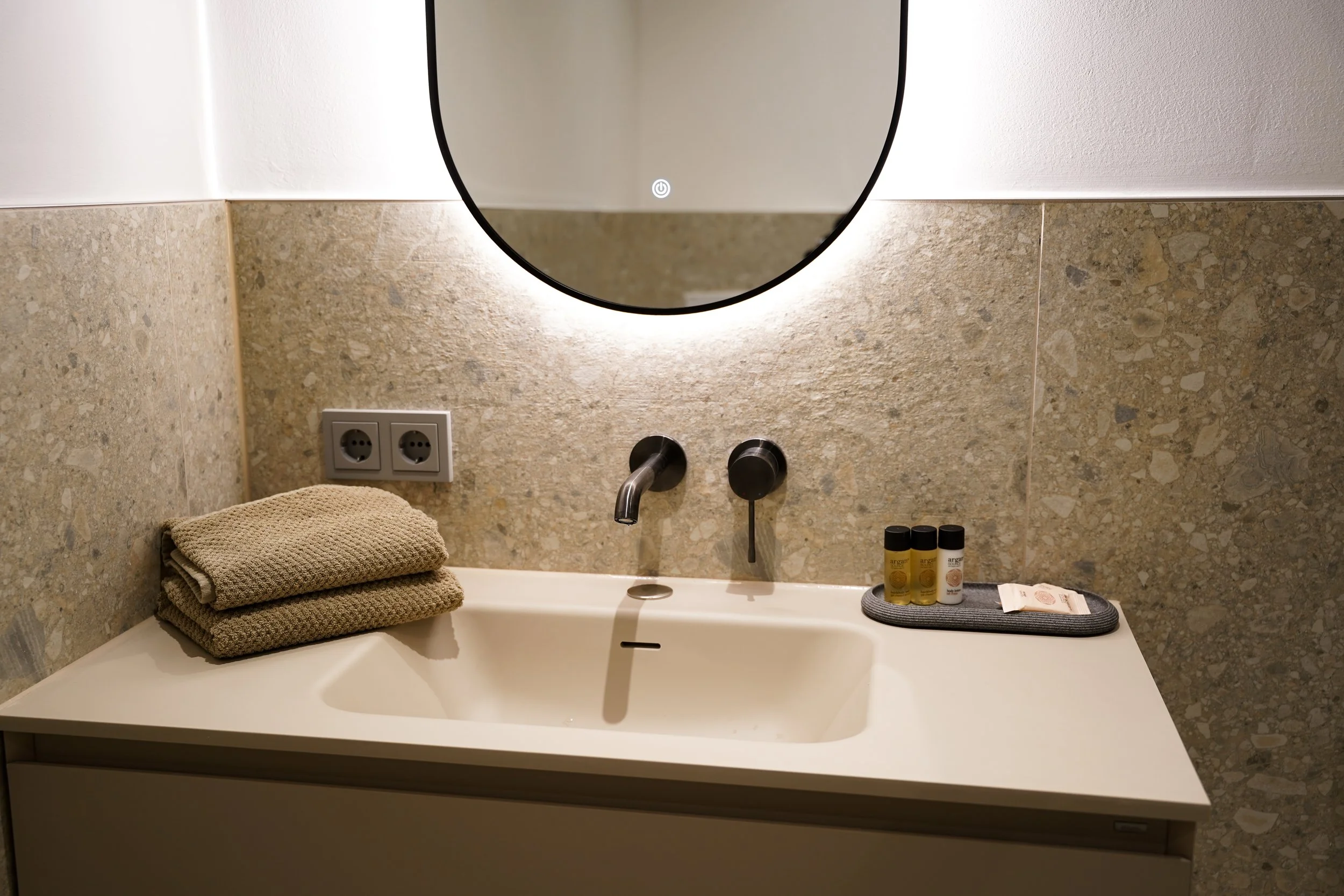 Waschbecken mit schwarzen Armaturen, beige Handtücher, drei kleine Pflegeprodukte, spiegelndes rundes Fenster, Wand mit Mosaikfliesen und Doppelsteckdose.