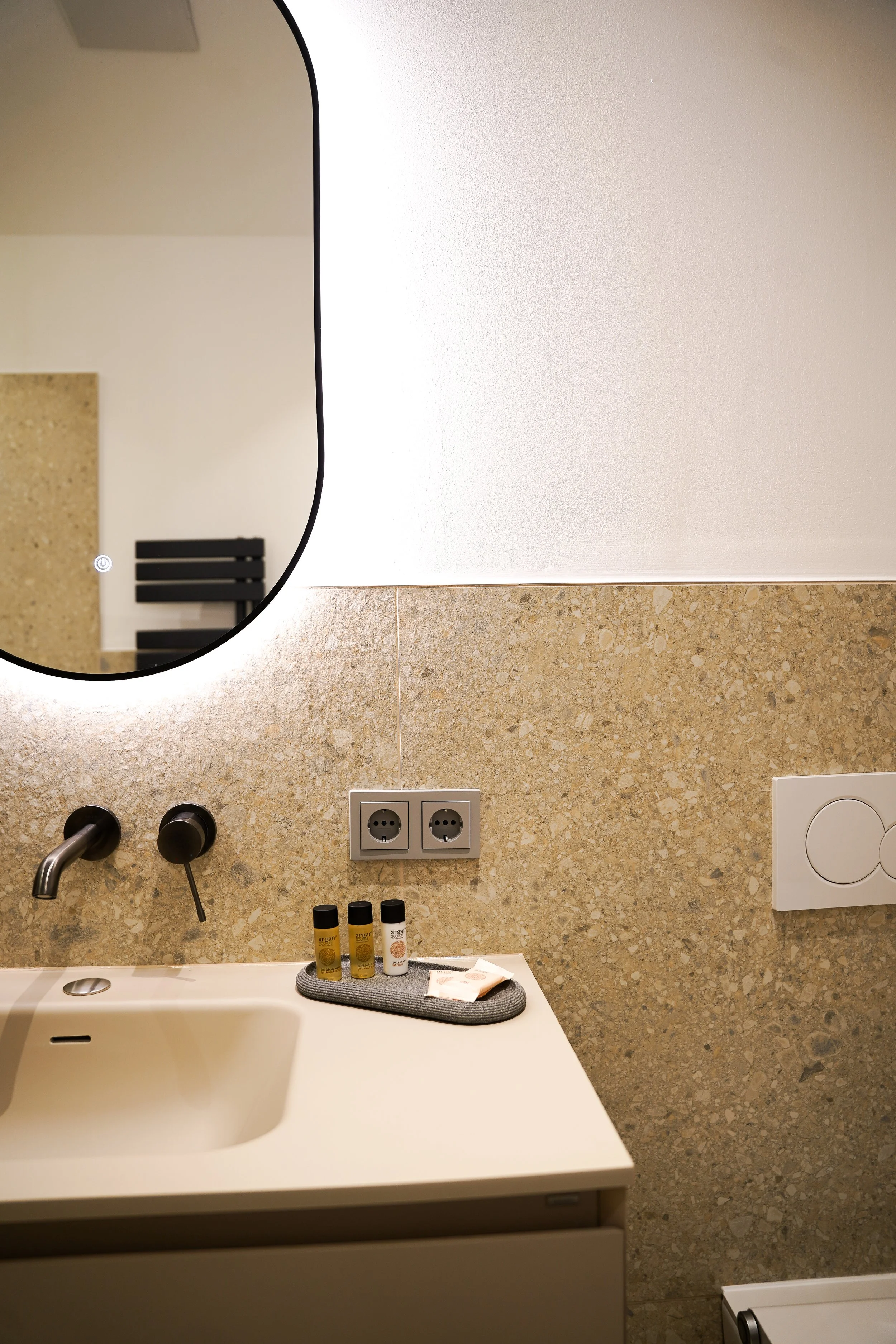 Waschbecken mit schwarzen Armaturen, Spiegel mit Beleuchtung, Steckdosen, kleine Flaschen und Seifenspender auf Waschtisch, beige Steinwand, weißer Wand