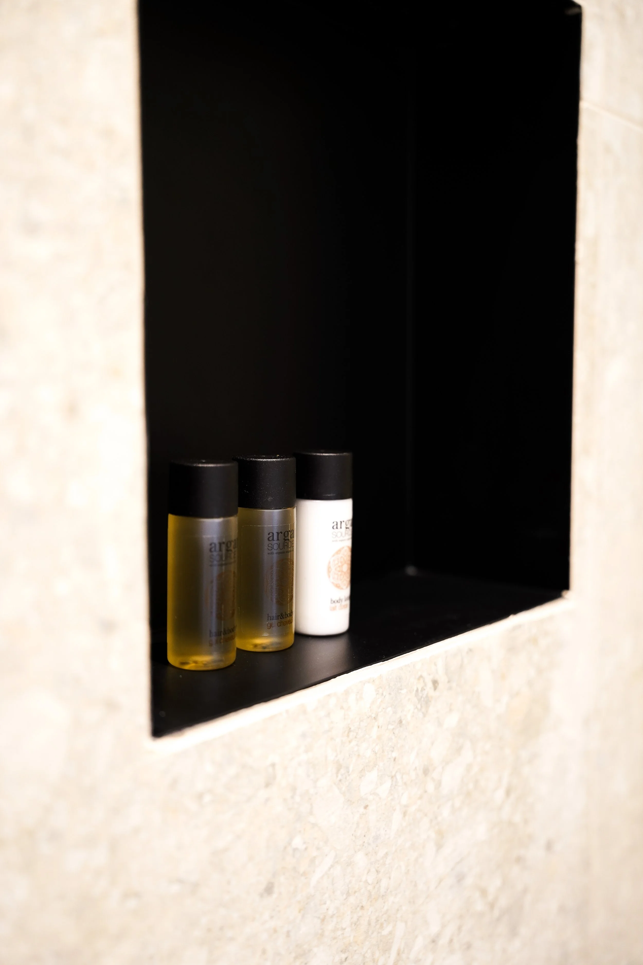 Kleine Flaschen mit Toilettenartikeln in einer schwarzen Nische, eingebettet in eine Wand aus hellen Steinen.