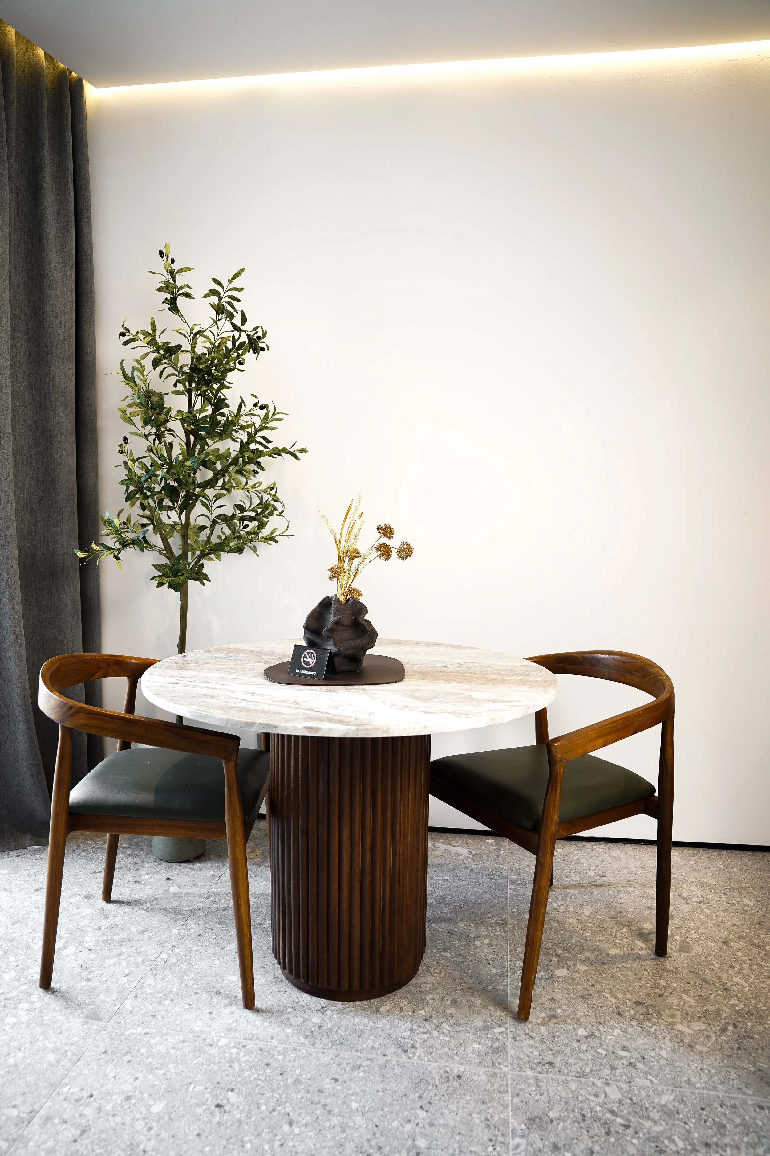 Ein runder Tisch mit weißer Marmorplatte, zwei holzgestühlte Stühle, eine dunkle Vase mit getrockneten Blumen, ein kleines Schild mit Durchgestrichenem Rauchzeichen, im Hintergrund eine große Topfpflanze an einer weißen Wand.