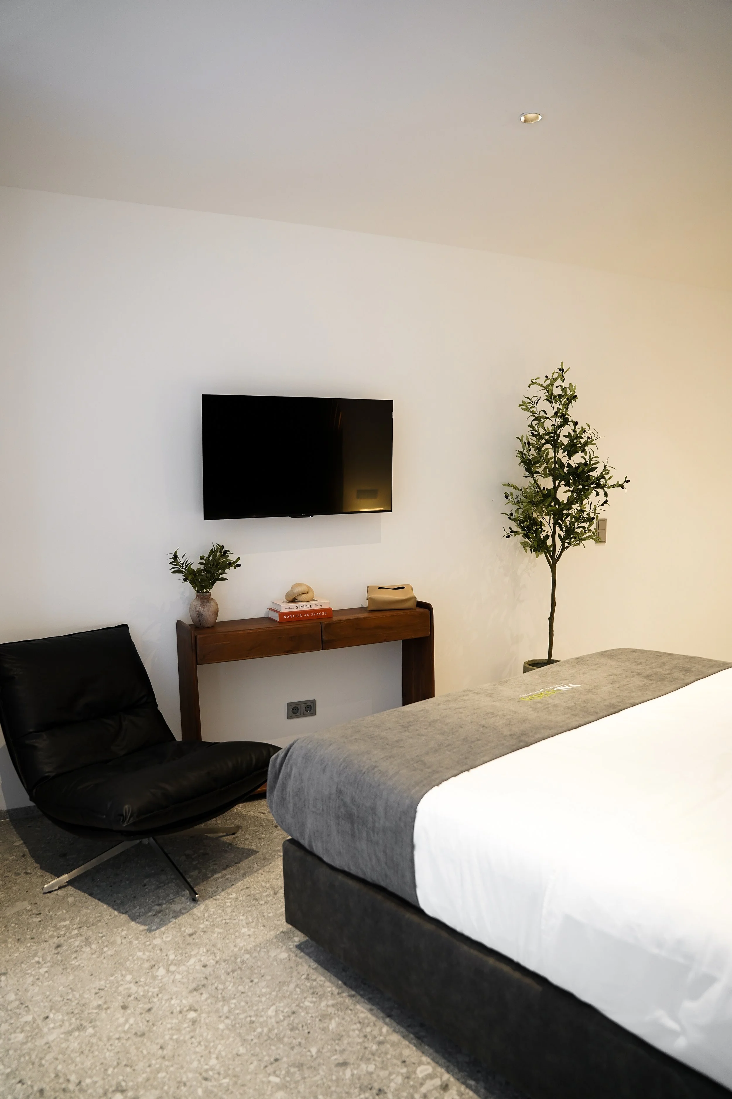 Modernes Schlafzimmer mit schwarz Lederstuhl, Wandfernseher, Holzkonsole, Zimmerpflanzen, und weißem Bett mit grauem Überwurf.