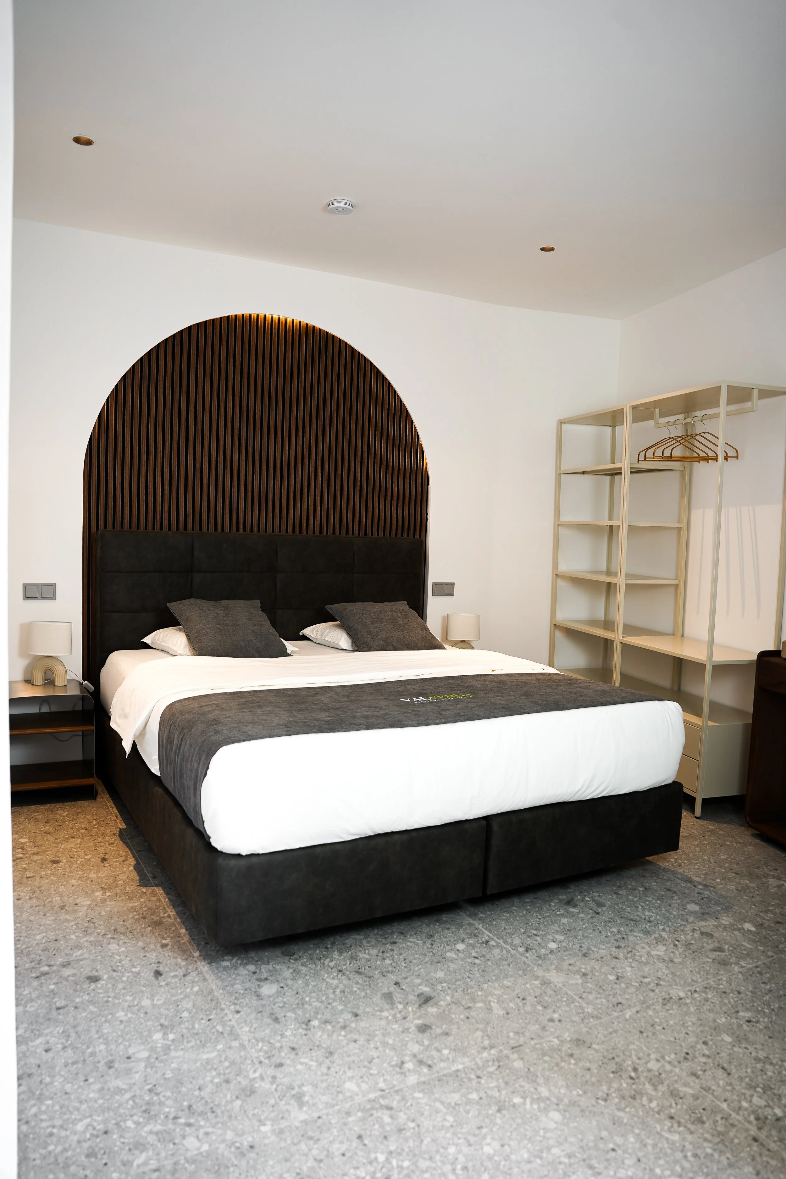 Modernes Schlafzimmer mit schwarzem Bett, grauen Kissen, weißen Wänden und offenem Kleiderschrank aus Holz.