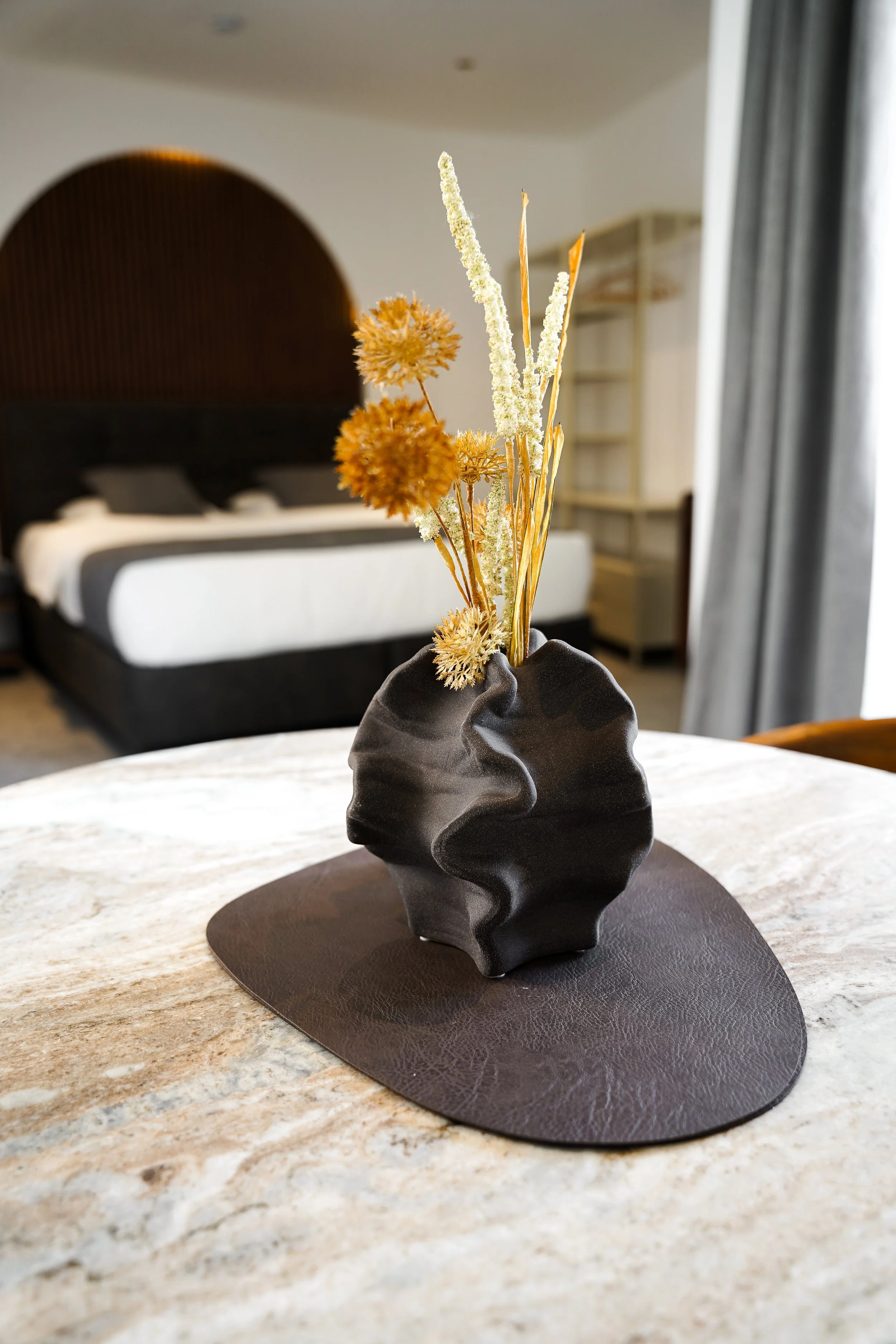 Kunstvolle schwarze Vase mit getrockneten Pflanzen auf einem Tisch, Schlafzimmer im Hintergrund.