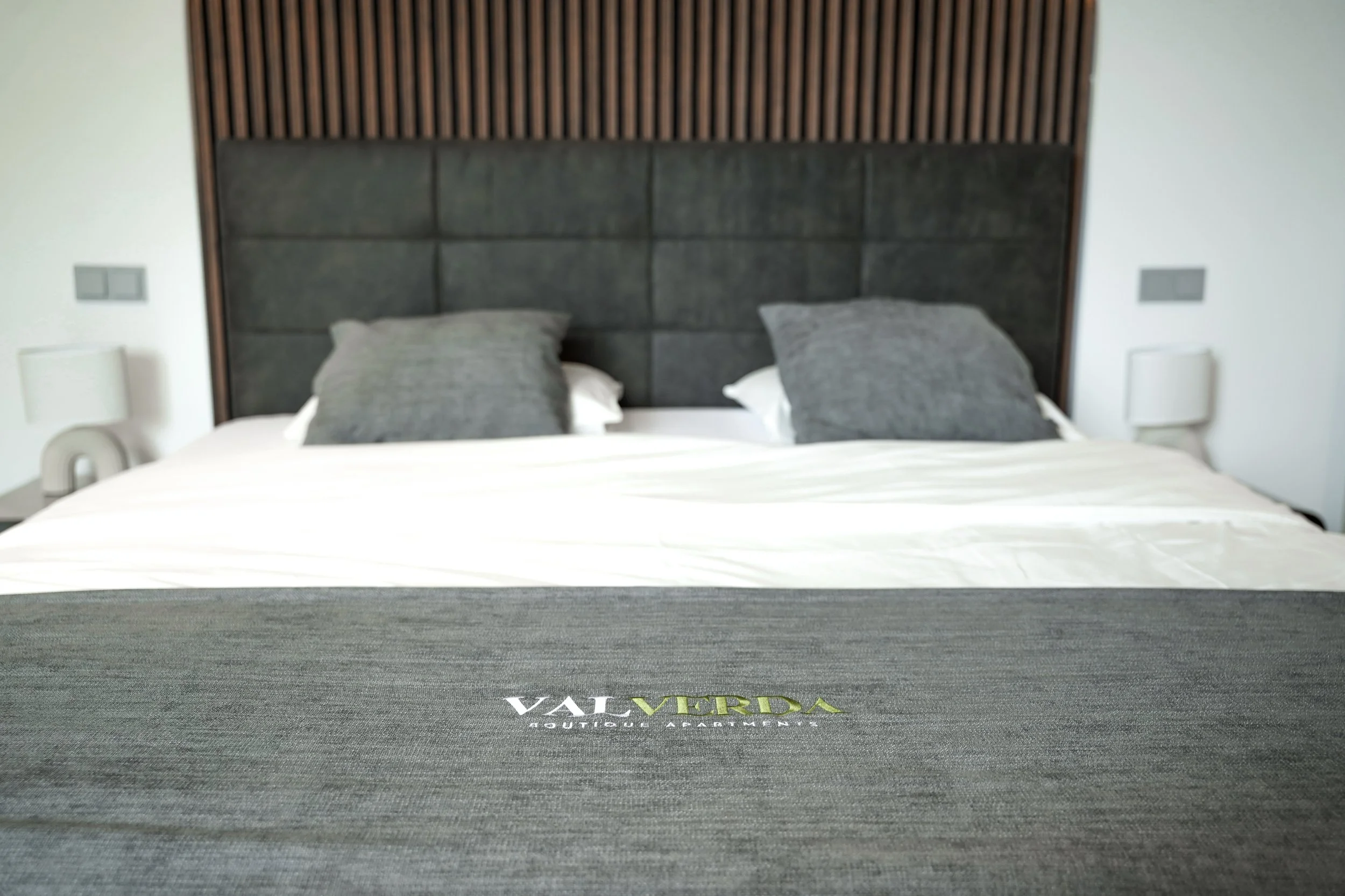 Bett mit grauem Kopfteil und weißer Bettdecke, zwei graue Kissen, schwarzer Tagesdecke mit weißer und grüner Schrift, graue Wandleuchten auf beiden Seiten, Holzverkleidung im Hintergrund.