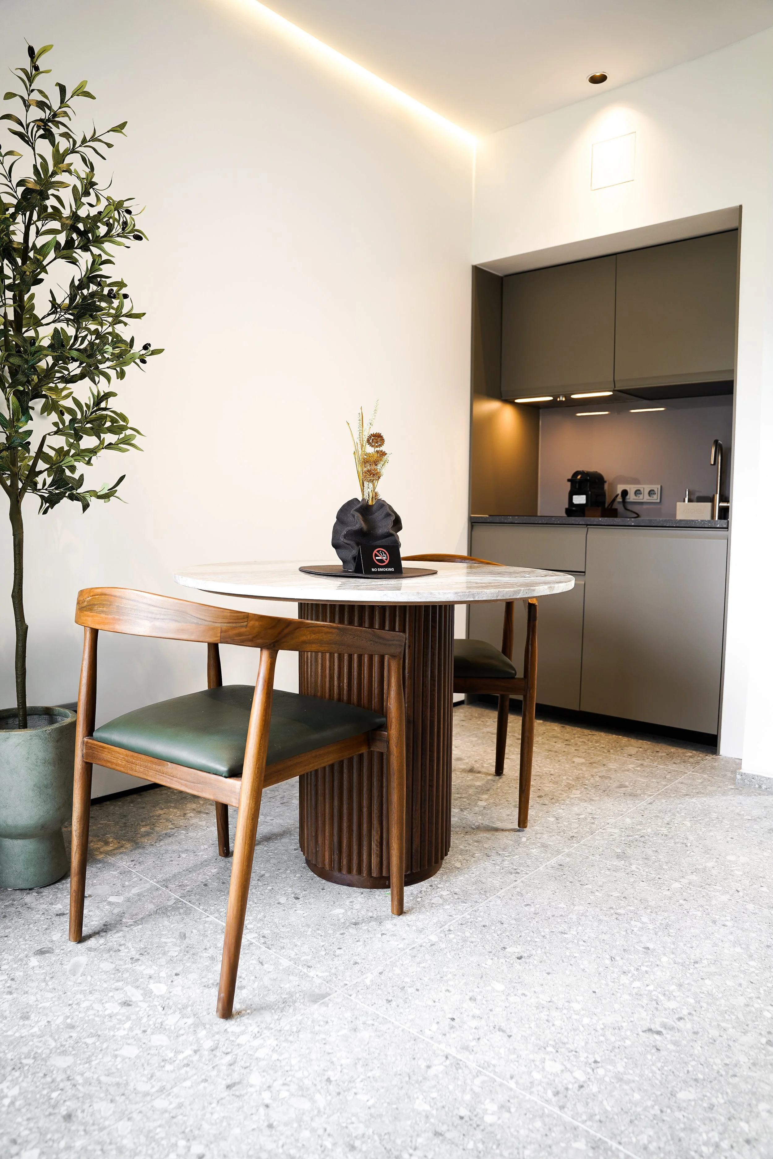 Ein kleiner Esstisch aus Marmor mit einer Vase und zwei Stühlen, eine Topfpflanze und ein offener Küchenbereich im Hintergrund.