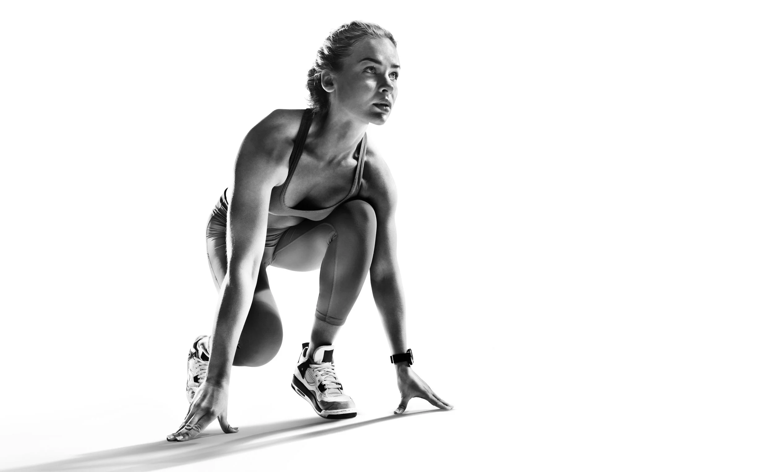 A woman in athletic gear crouches in a starting position, ready for a race, in a black and white studio shot.