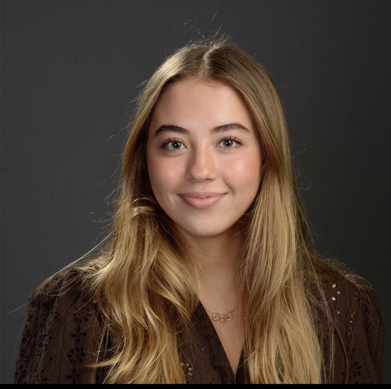 Portrait of a young woman with blonde hair, blue eyes, and a gentle smile, wearing a brown top and a small silver bow necklace against a dark gray background.