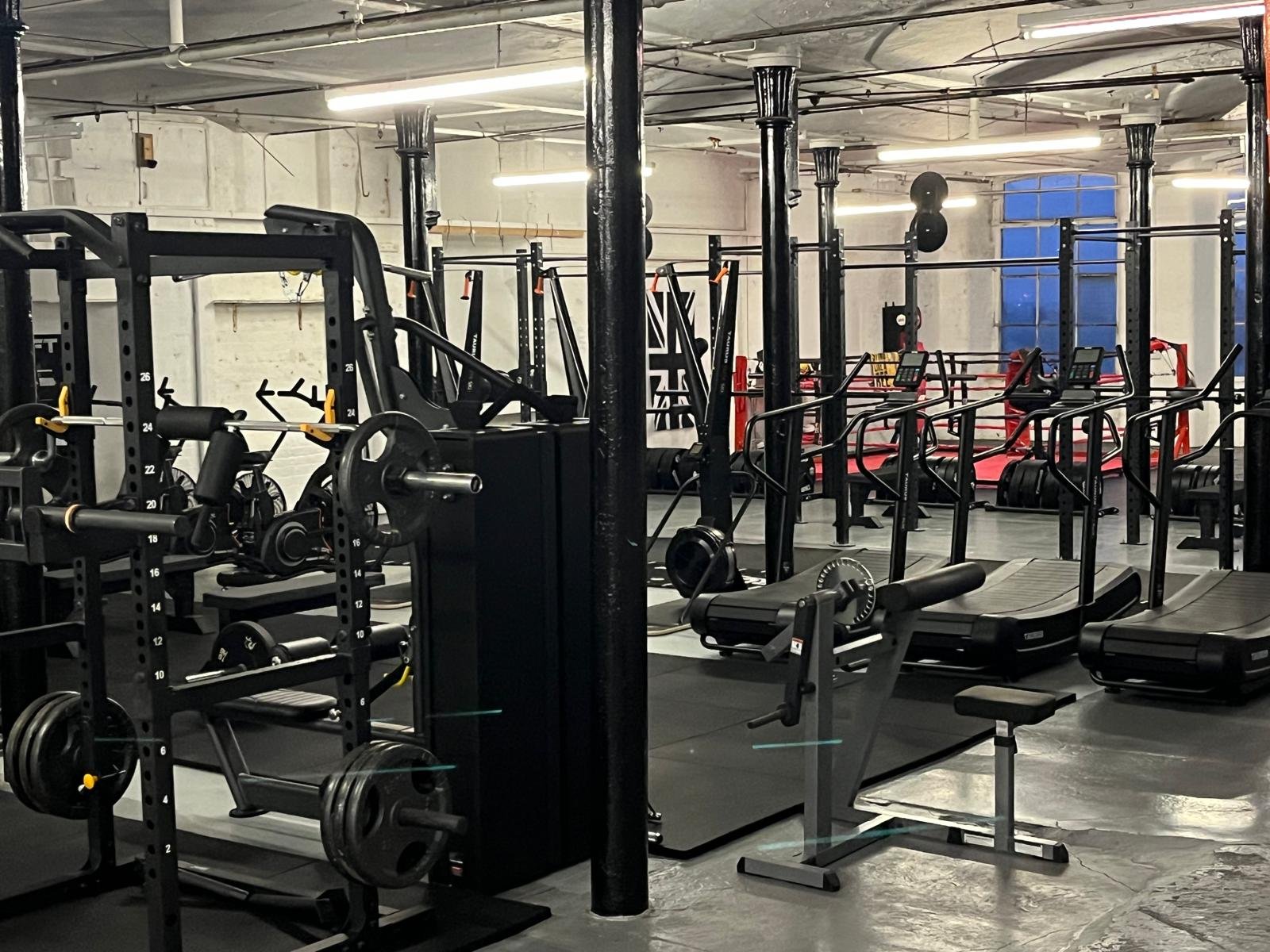 Empty commercial gym with various workout equipment including treadmills, weight racks, and benches, in an industrial-style space with white brick walls and large windows.