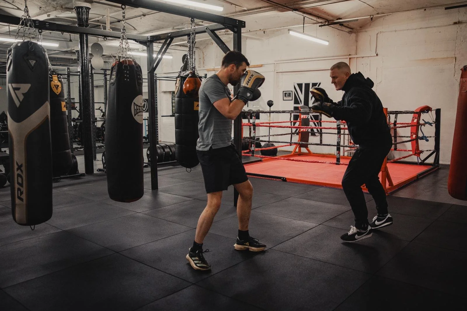 Two men practicing boxing in a gym, wearing boxing gloves, near punching bags and a boxing ring.