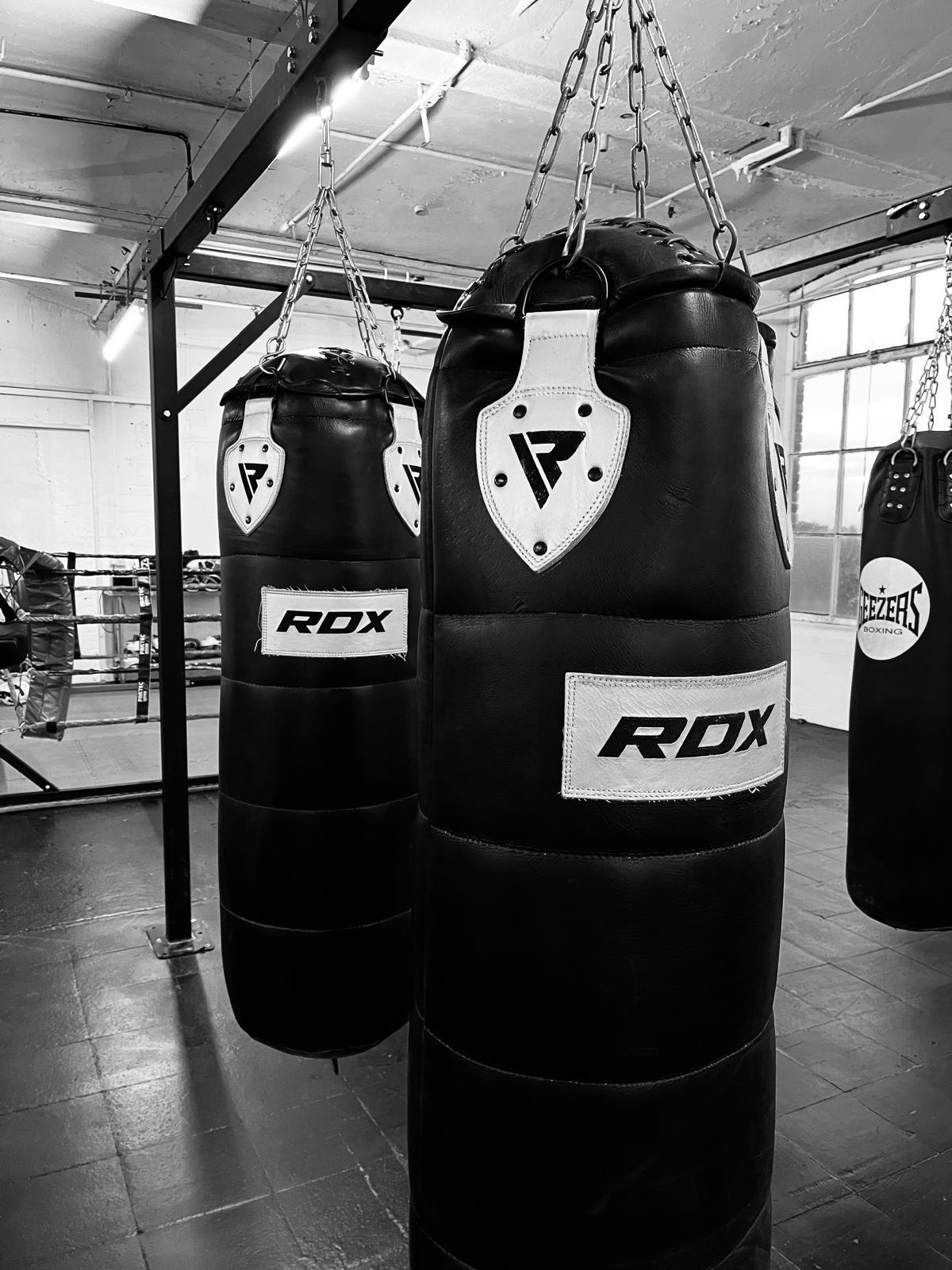 Two black punching bags hanging in a boxing gym, with a boxing ring and a person training in the background, in black and white.