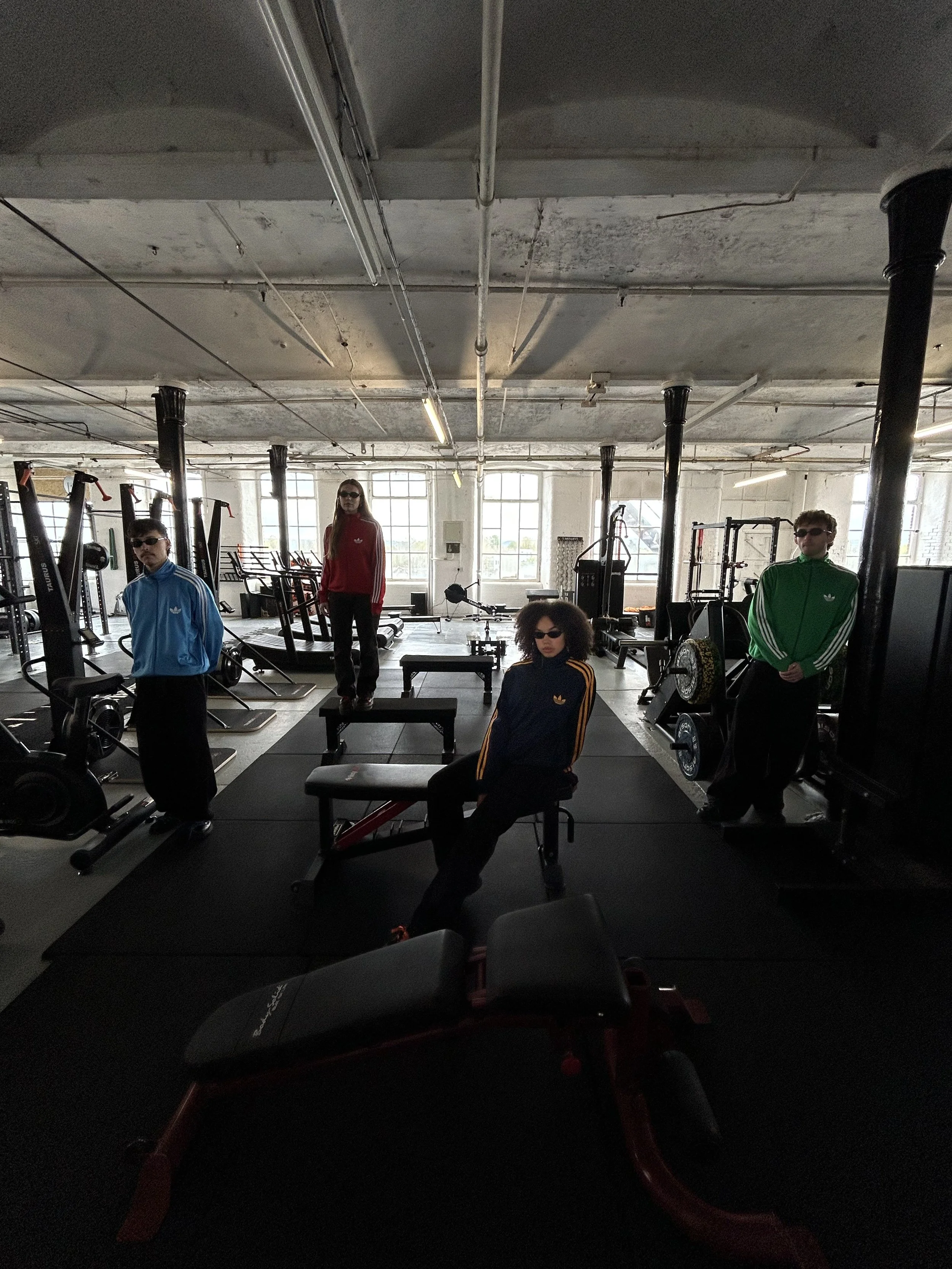 People in a gym with exercise equipment, large windows, and natural light, wearing colorful tracksuits, some with sunglasses.