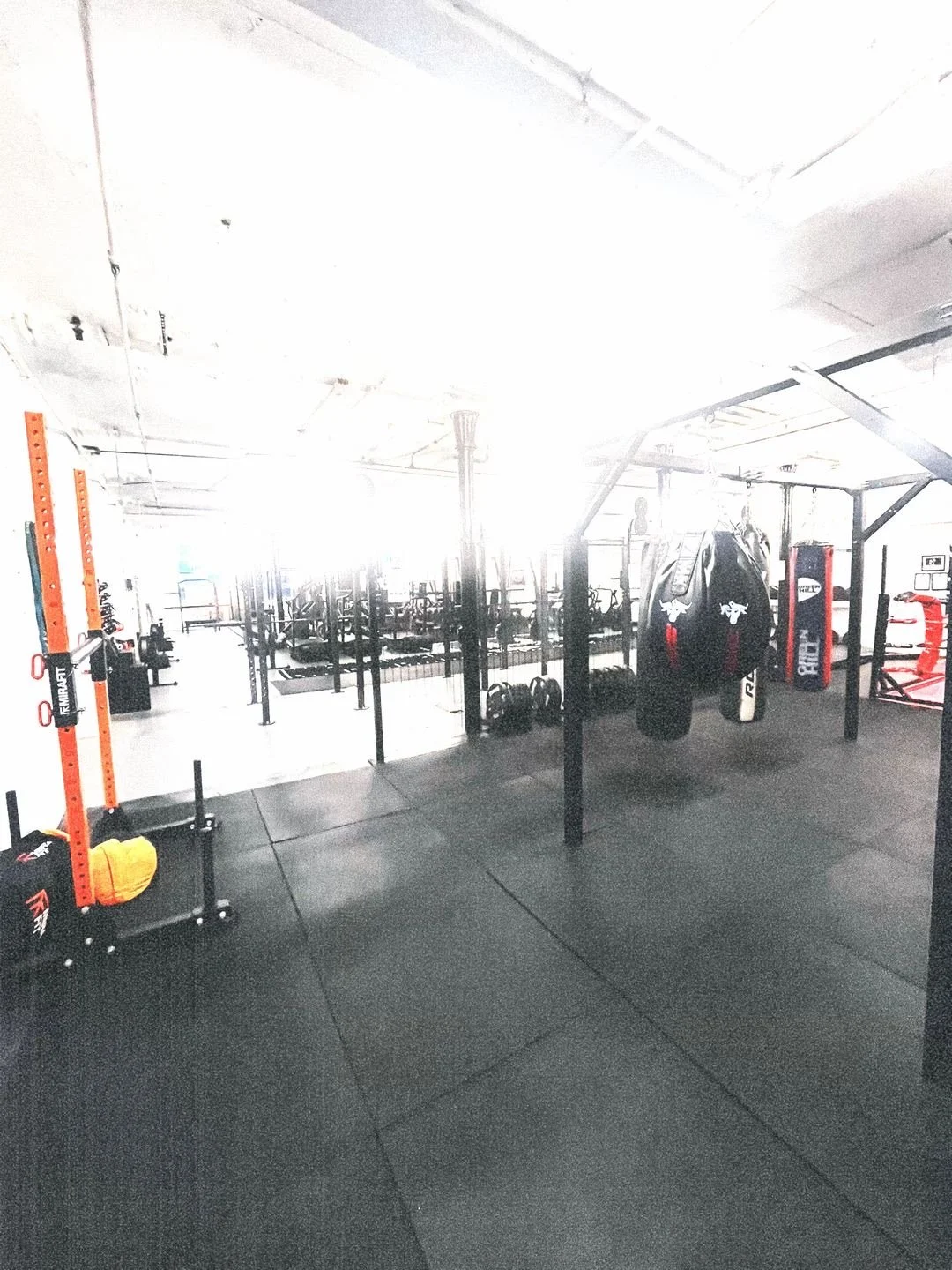 Empty gym with black rubber flooring, punching bags, weightlifting equipment, and open workout area.