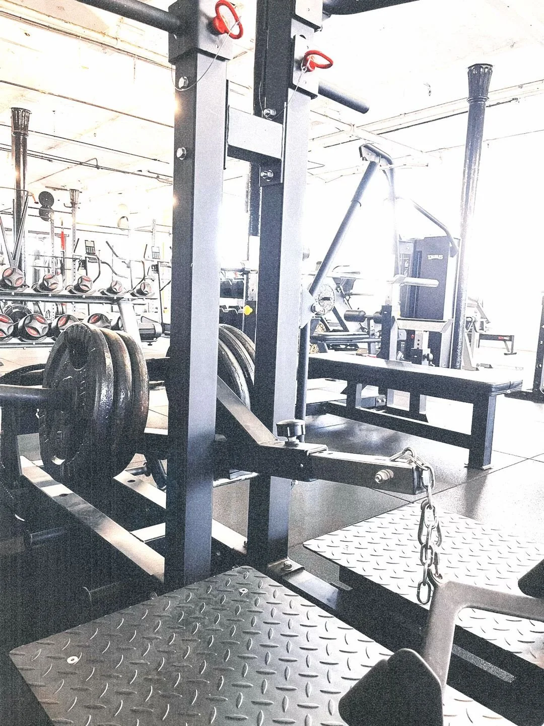 A gym with weight lifting equipment, including a squat rack with weights, a bench press, and other strength training machines and free weights in the background.