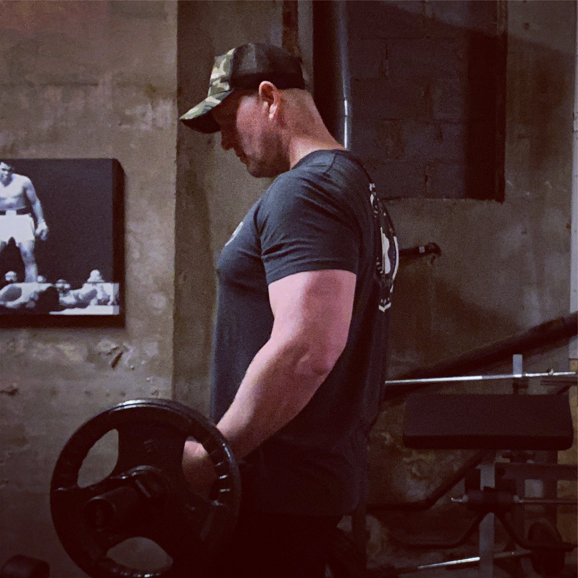 A man lifting a dumbbell in a gym with exposed brick walls and a framed picture of a tank in the background.