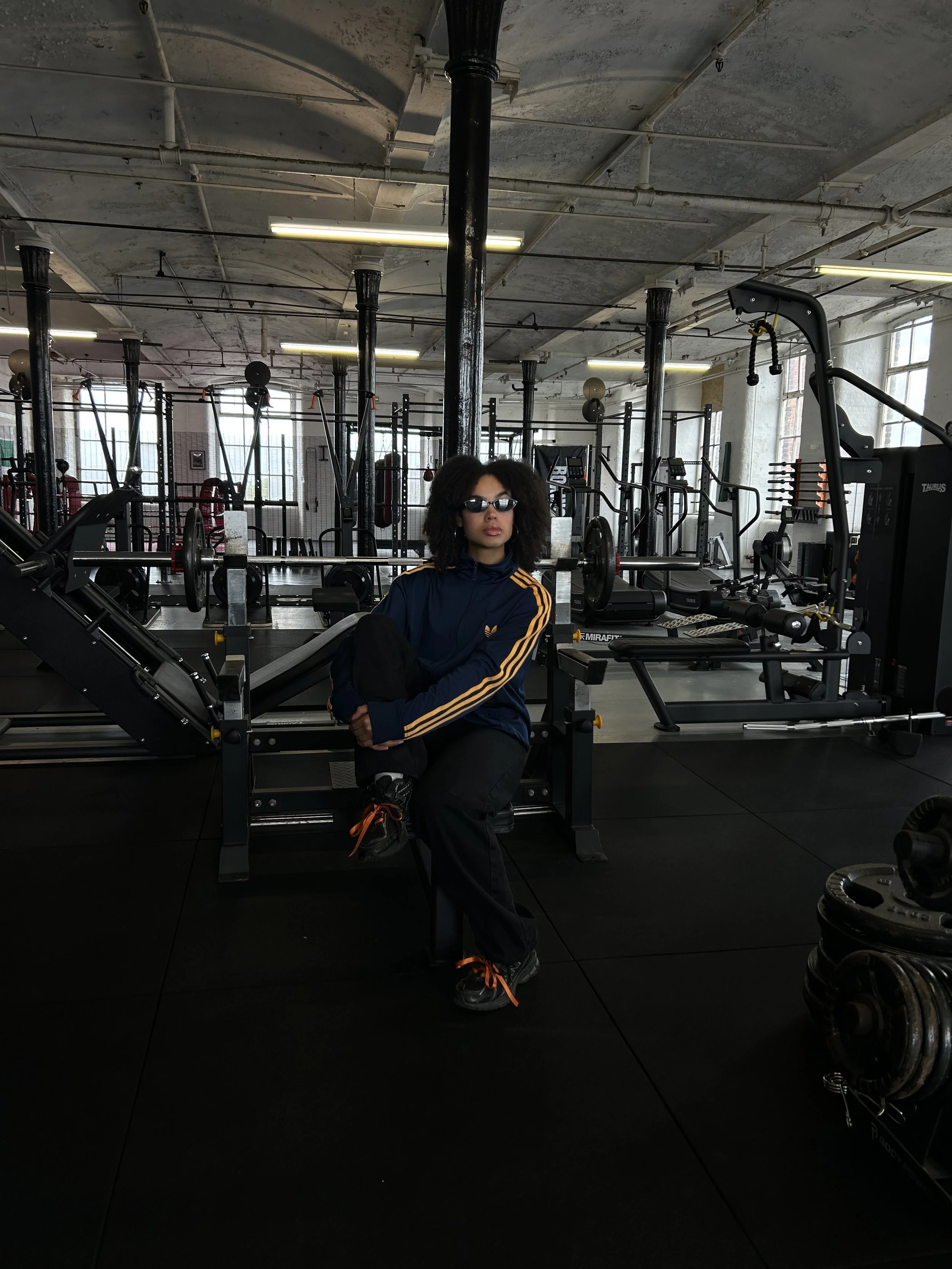 A woman with curly hair wearing sunglasses and a navy blue Adidas tracksuit with gold stripes sits on a weightlifting bench inside a spacious gym with large windows, black pillars, and various gym equipment in the background.