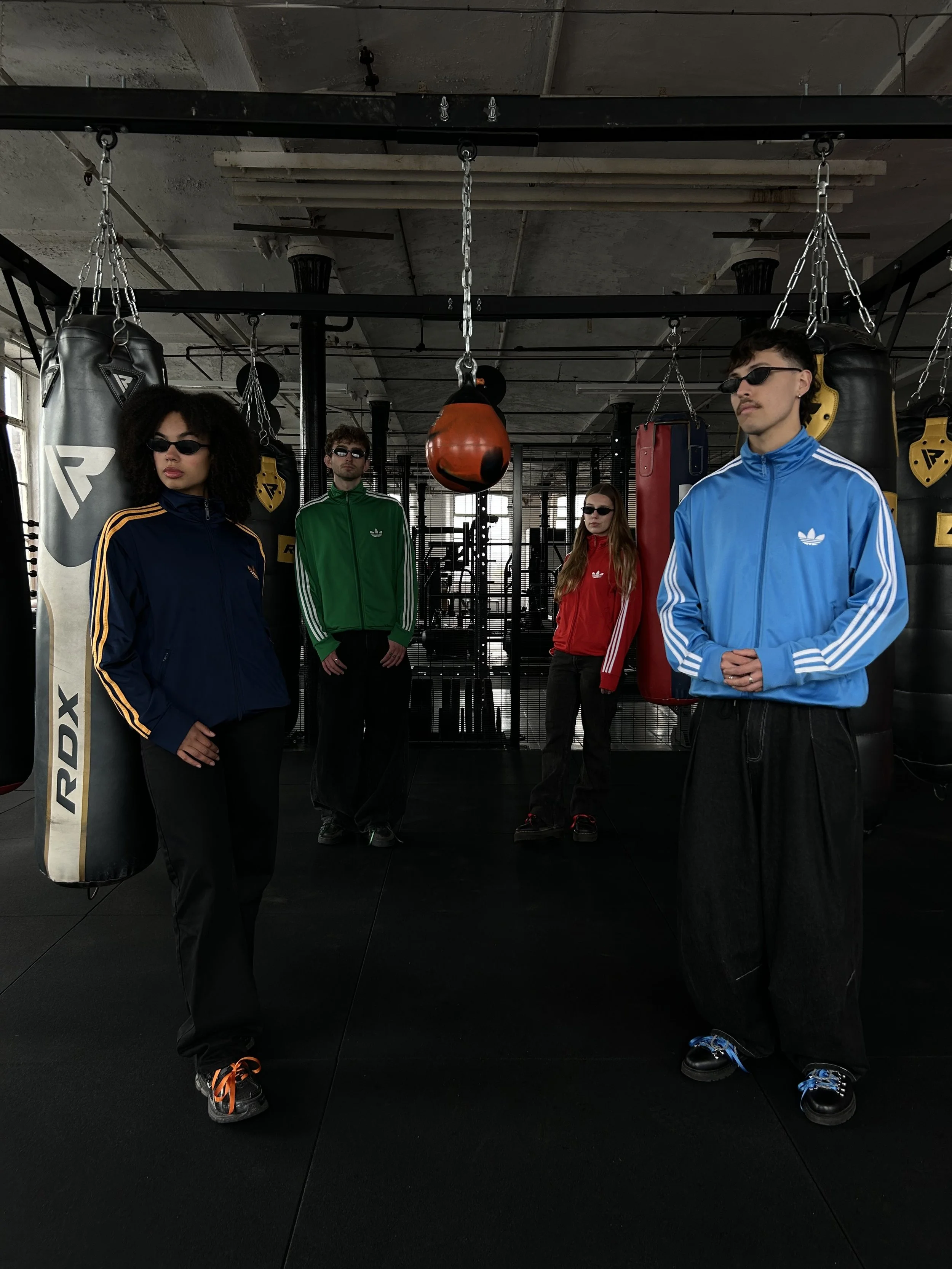 Four individuals standing in a gym with punching bags, dressed in colorful athletic jackets and sunglasses.