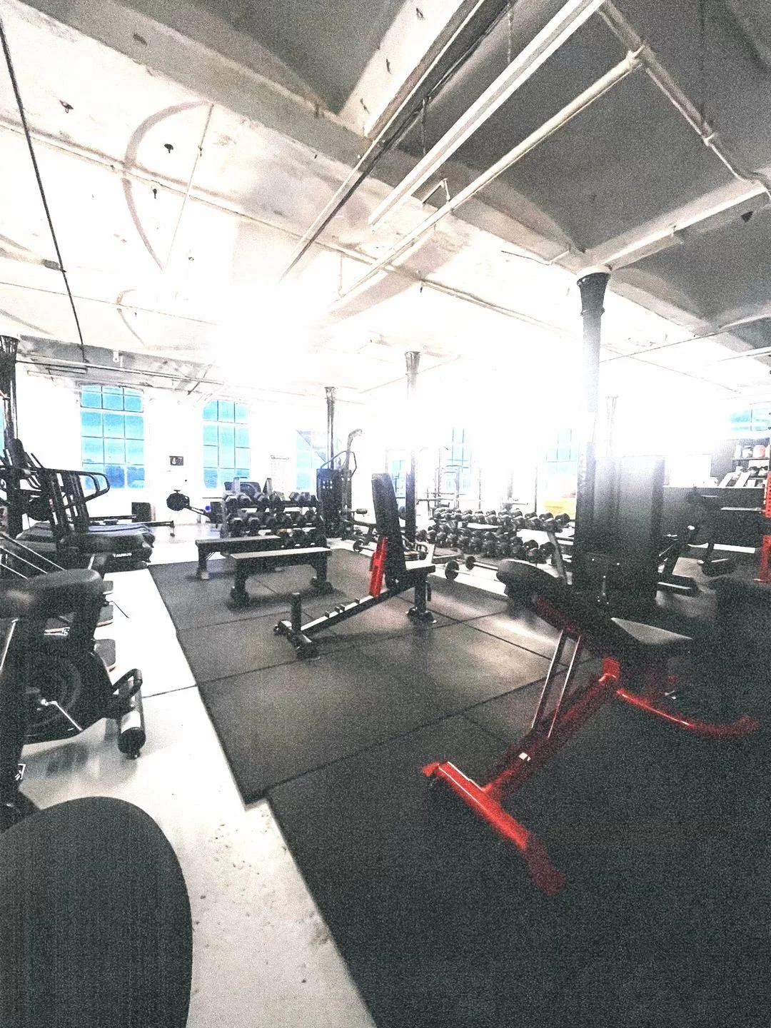 Interior of a well-lit gym with various workout equipment, weights, and benches on black rubber flooring, and large windows allowing natural light.
