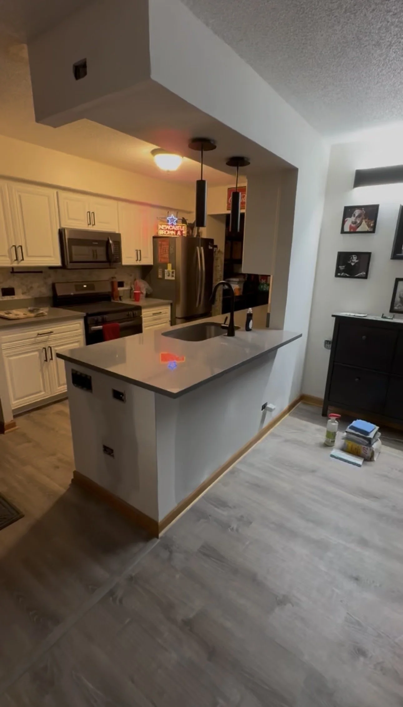 Photo of a modern kitchen with white cabinets, stainless steel appliances, a kitchen island with a black sink, and decorative framed photos on the wall. There are pendant lights hanging above the island, and some items on the countertop and floor.
