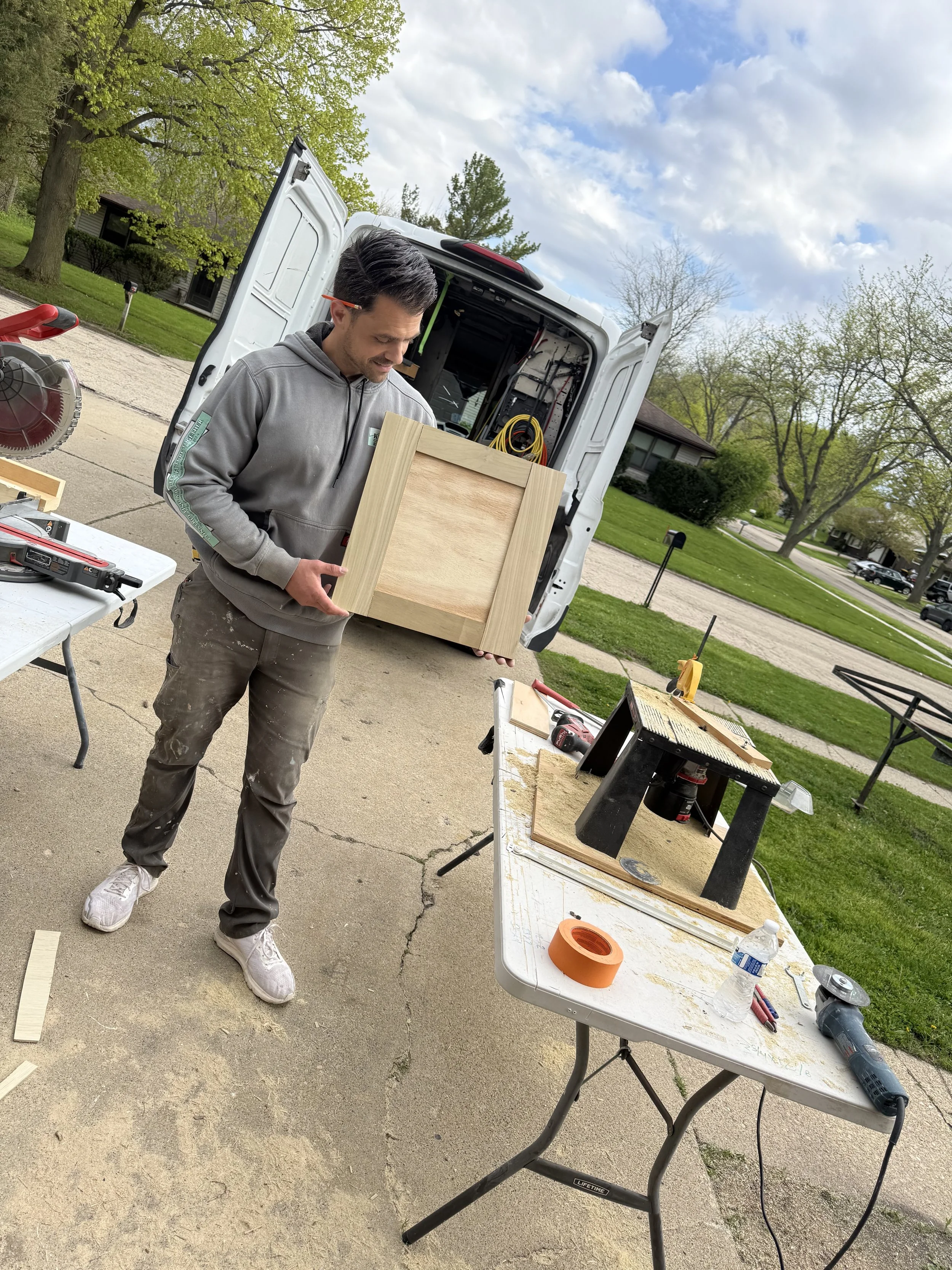 A man in a gray hoodie holds a wooden piece in front of an open van with construction tools inside. There are worktables with tools and supplies outdoors on a driveway, with trees, a house, and a cloudy sky in the background.