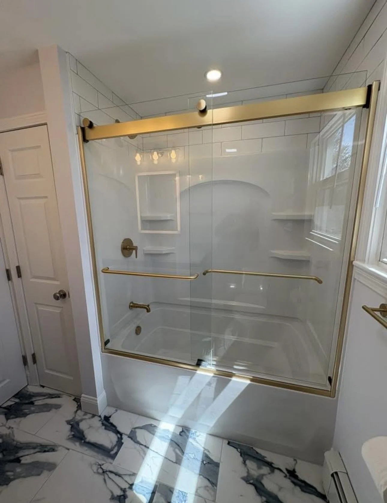 Bathroom with a walk-in shower featuring glass doors with gold trim, white tiled walls, built-in shower shelves, and gold grab bars. Marble flooring and a window providing natural light.