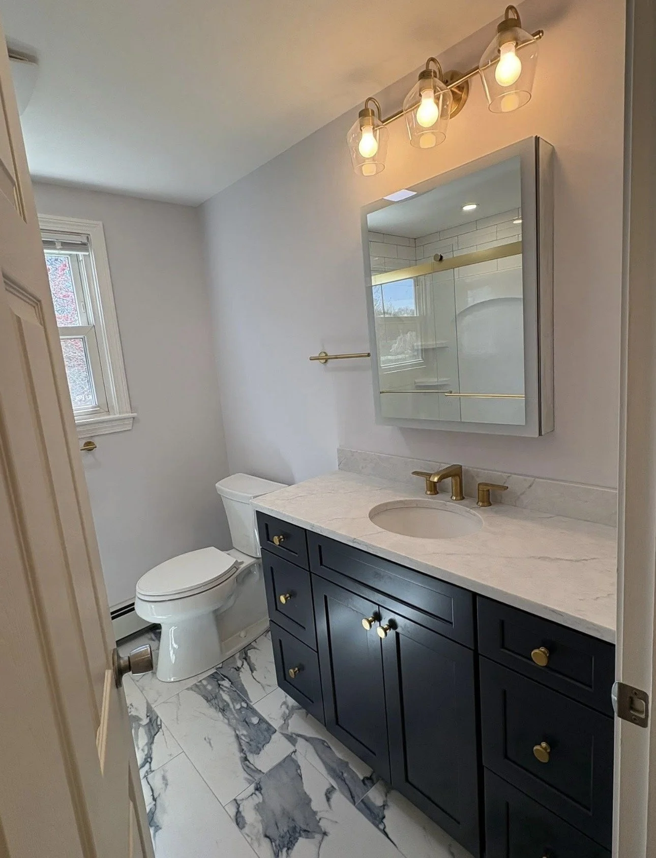 Modern bathroom with marble floor, navy blue vanity with gold hardware, white marble countertop, under-mount sink, and gold faucet. Above the vanity is a mirrored cabinet, and above that are three light bulbs in clear glass fixtures. To the left is a white toilet next to a window with white trim. The walls are painted light gray, and the bathroom has a minimalistic, elegant design.