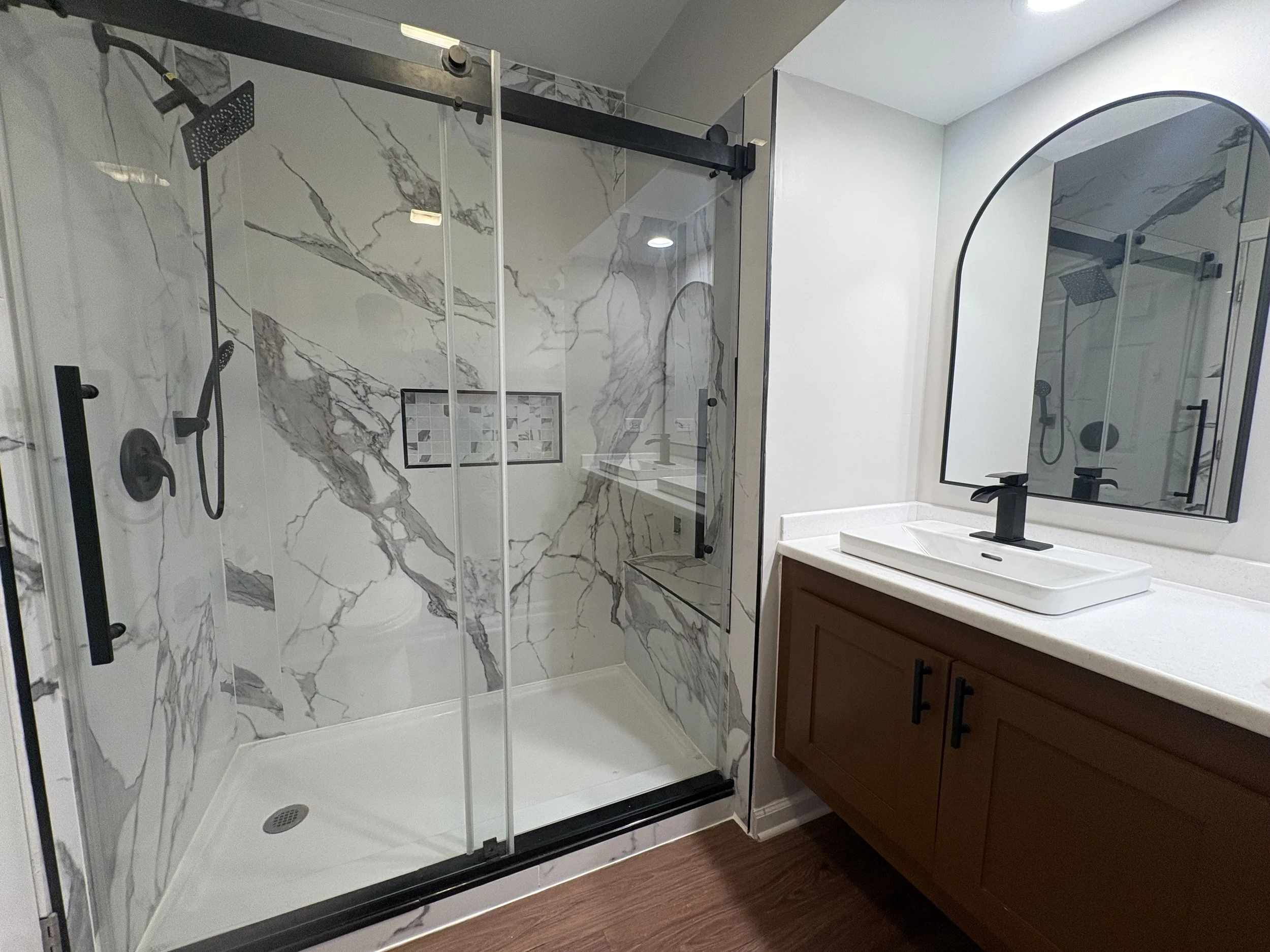 A bathroom with a walk-in shower featuring white marble walls with gray veining, a glass shower door with black hardware, and a built-in tiled niche. There is a modern white vanity with a black faucet and a large arched mirror above it. The flooring is wood-like.
