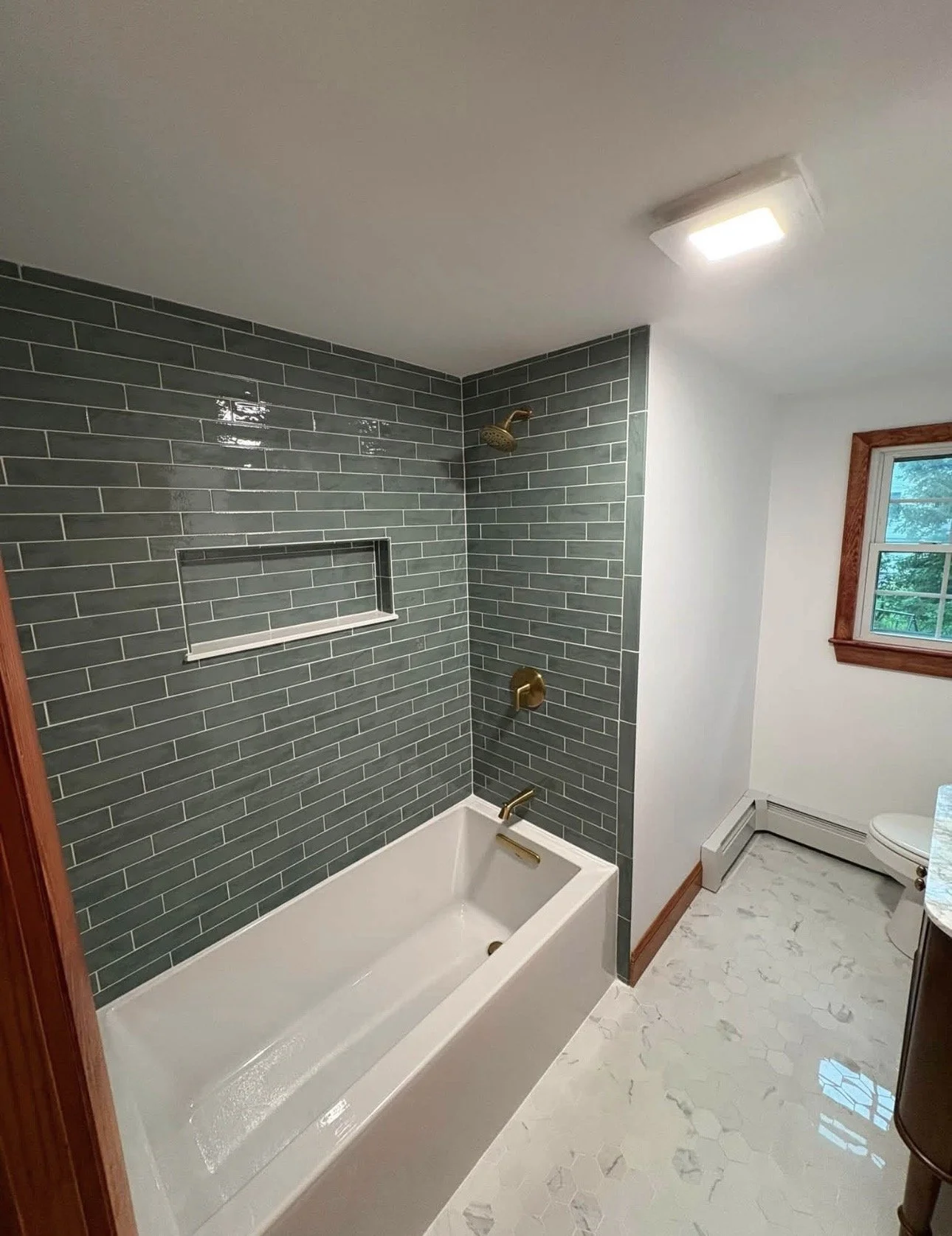 Bathroom with white bathtub, green tiled shower wall, brass fixtures, window with wooden trim, white marble floor with gray veining, and white walls.