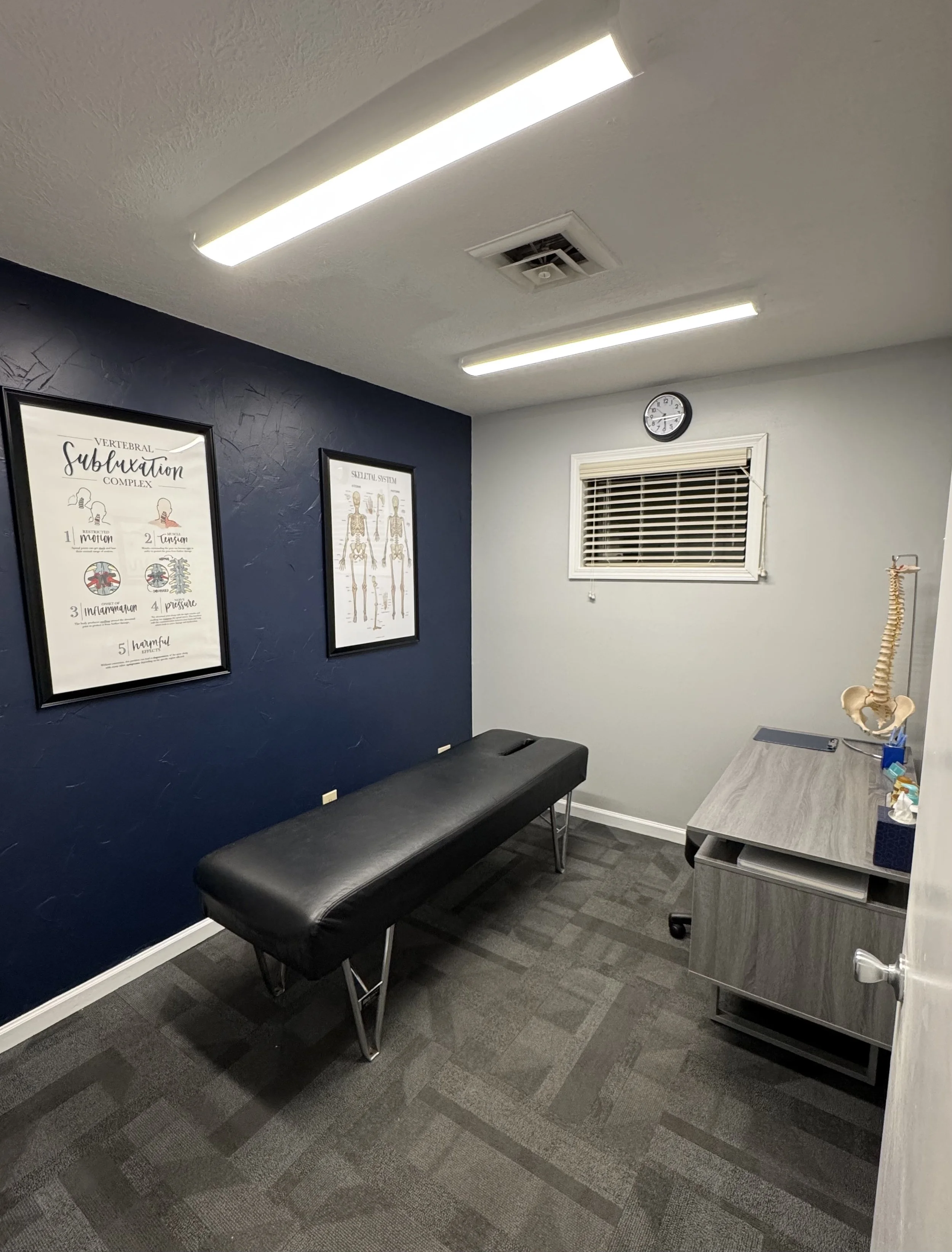 Medical examination room with a black examination table, framed anatomical posters on a blue accent wall, a wall clock, a window with blinds, and a desk with a model of a human spine.
