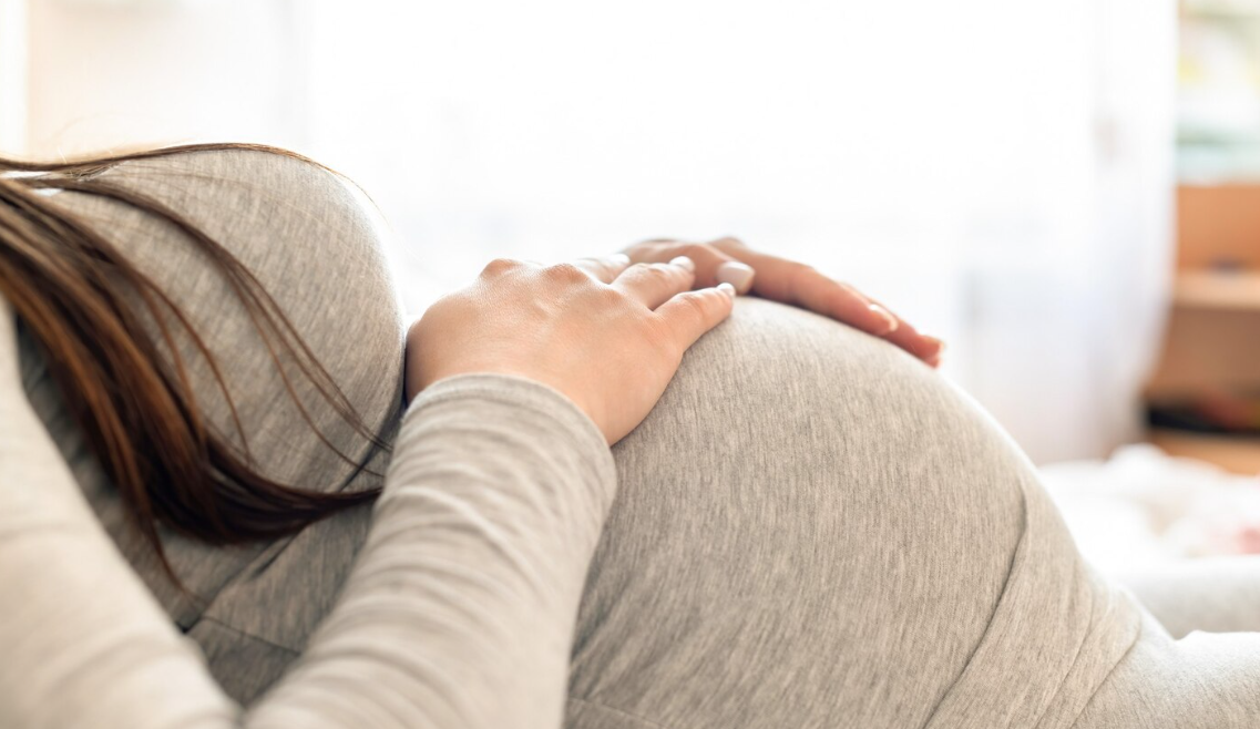 A person resting their hand on their pregnant belly, which is covered by a light gray shirt, sitting on a beige sofa.