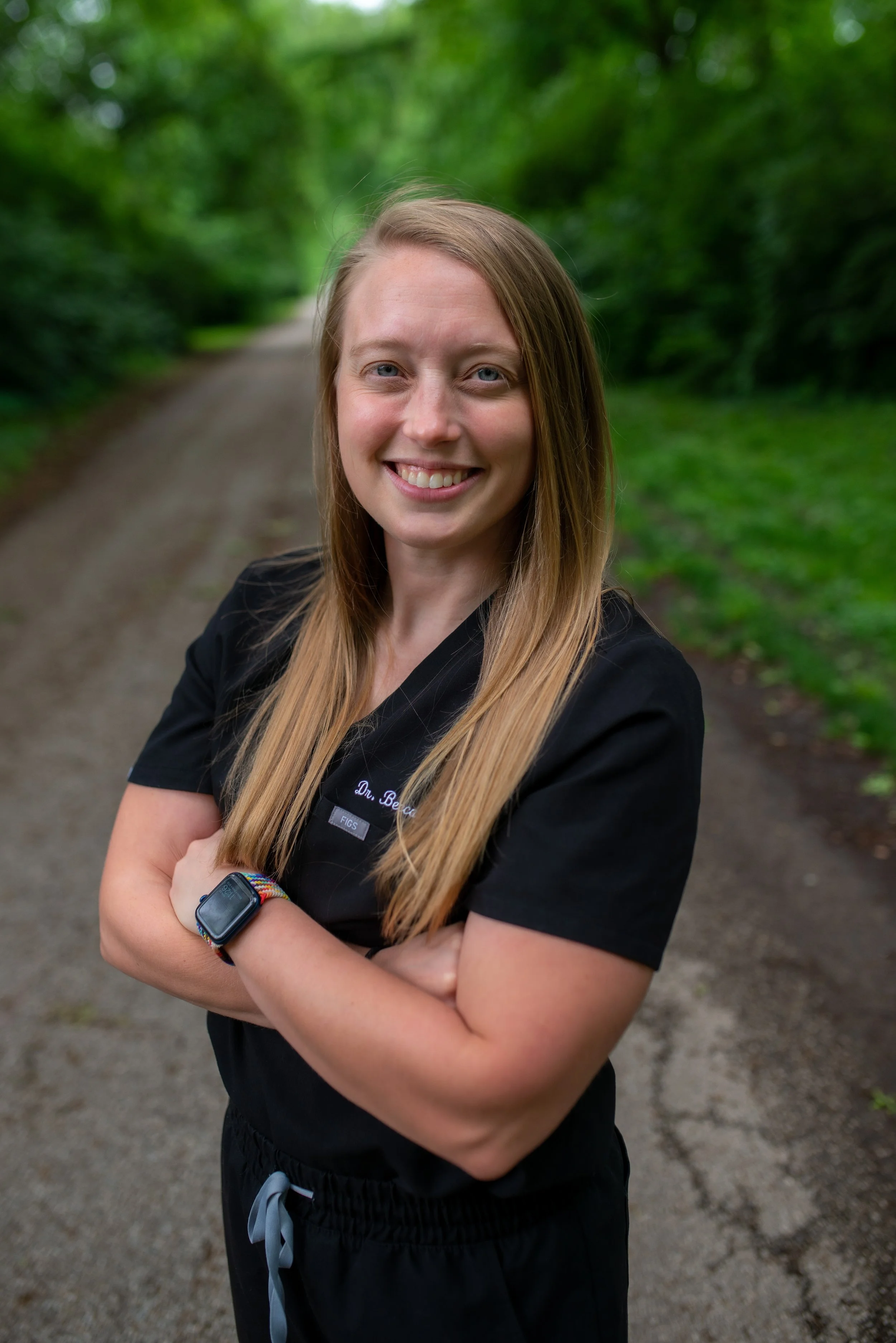 A woman with long blonde hair, smiling, standing outdoors on a dirt path surrounded by green trees, wearing a black athletic top and a smartwatch.