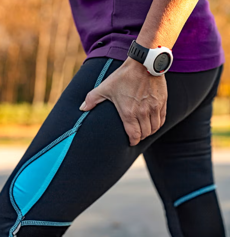 Close-up of a person's hand resting on their thigh, wearing a black watch with a black and white band, outdoors with blurred trees in the background