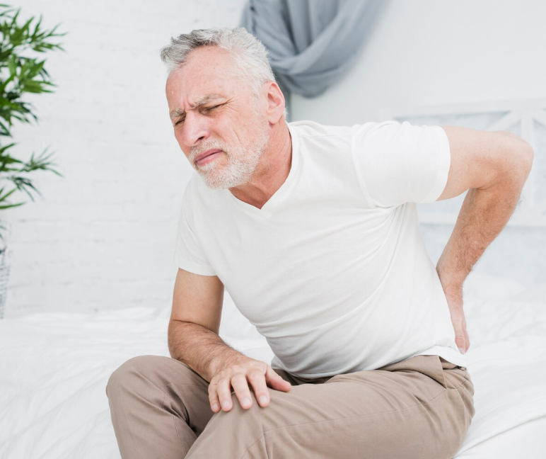 A woman with short blonde hair and wearing an orange sleeveless top and beige pants sits on a beige seat, writing in a notebook. She is in a room with sunlight streaming through a window, creating shadows on the wall behind her. A large green plant and a framed abstract artwork are also visible.