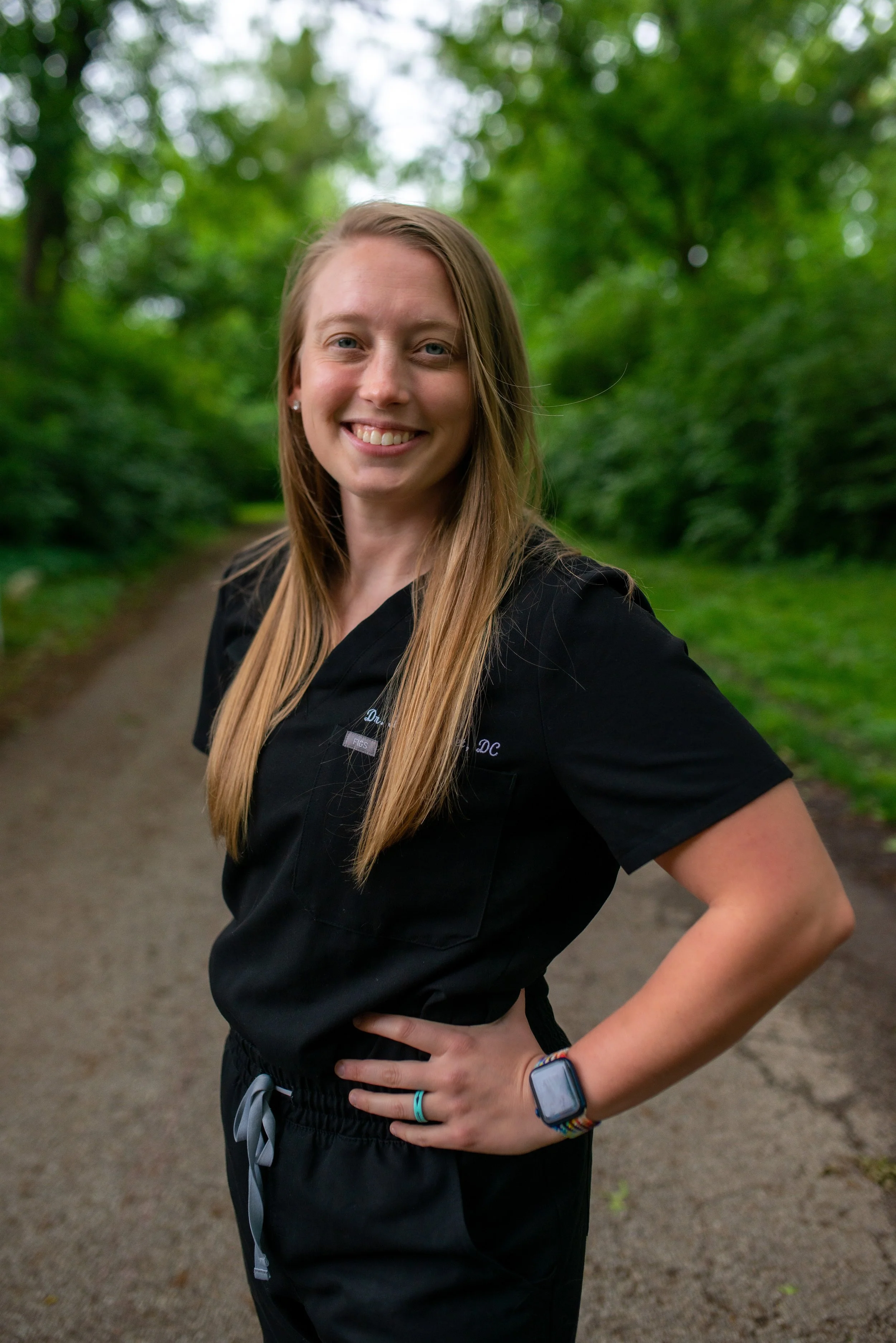 A woman with long blonde hair smiling outdoors on a path surrounded by green trees.