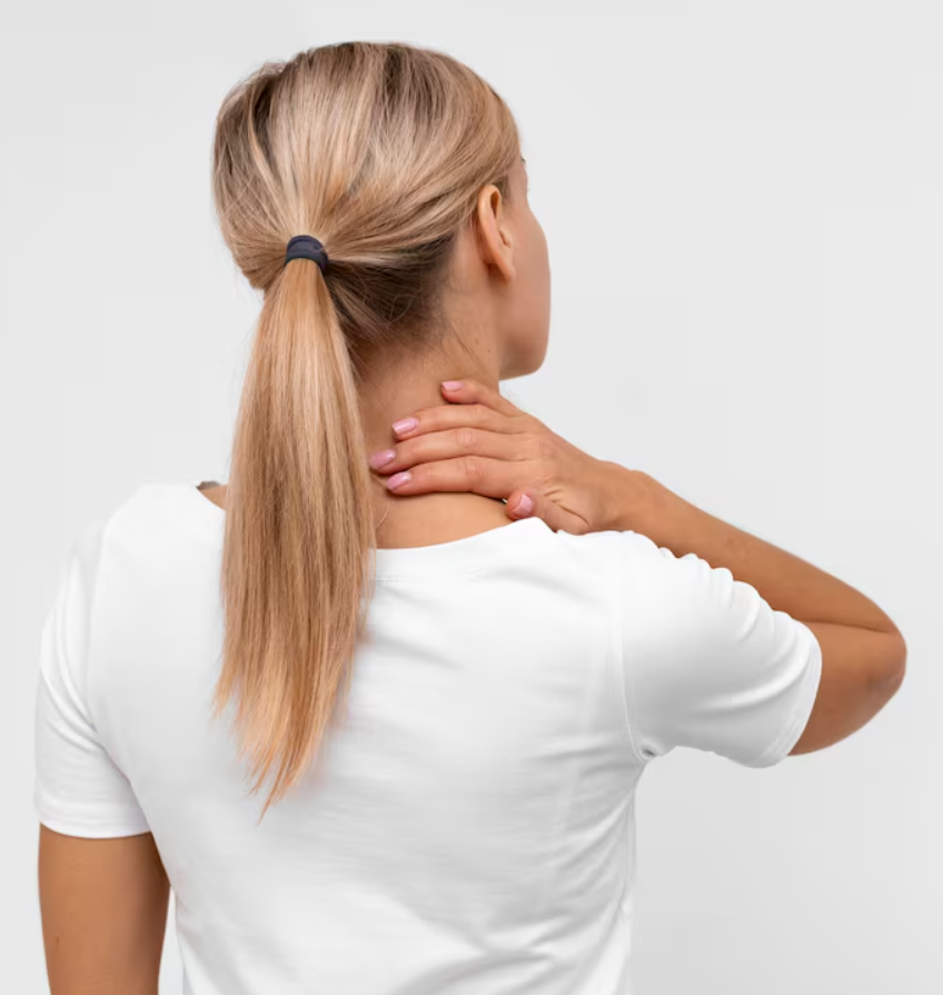 A woman with blonde hair tied in a ponytail experiencing neck pain, touching her neck with her right hand, against a plain white background.
