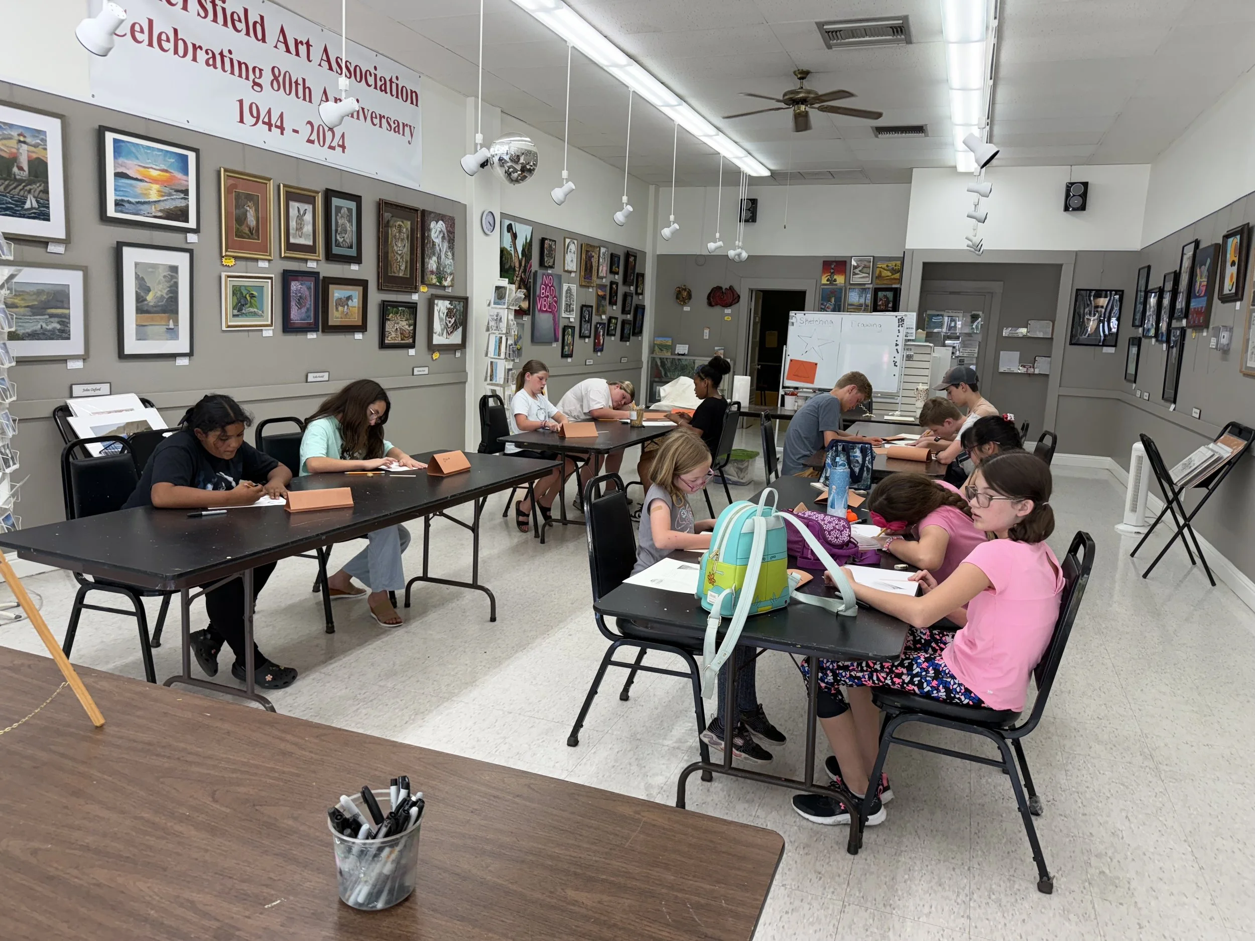 Children sitting at tables in an art classroom, focused on drawing and writing, with artwork displayed on the walls and a whiteboard in the background.