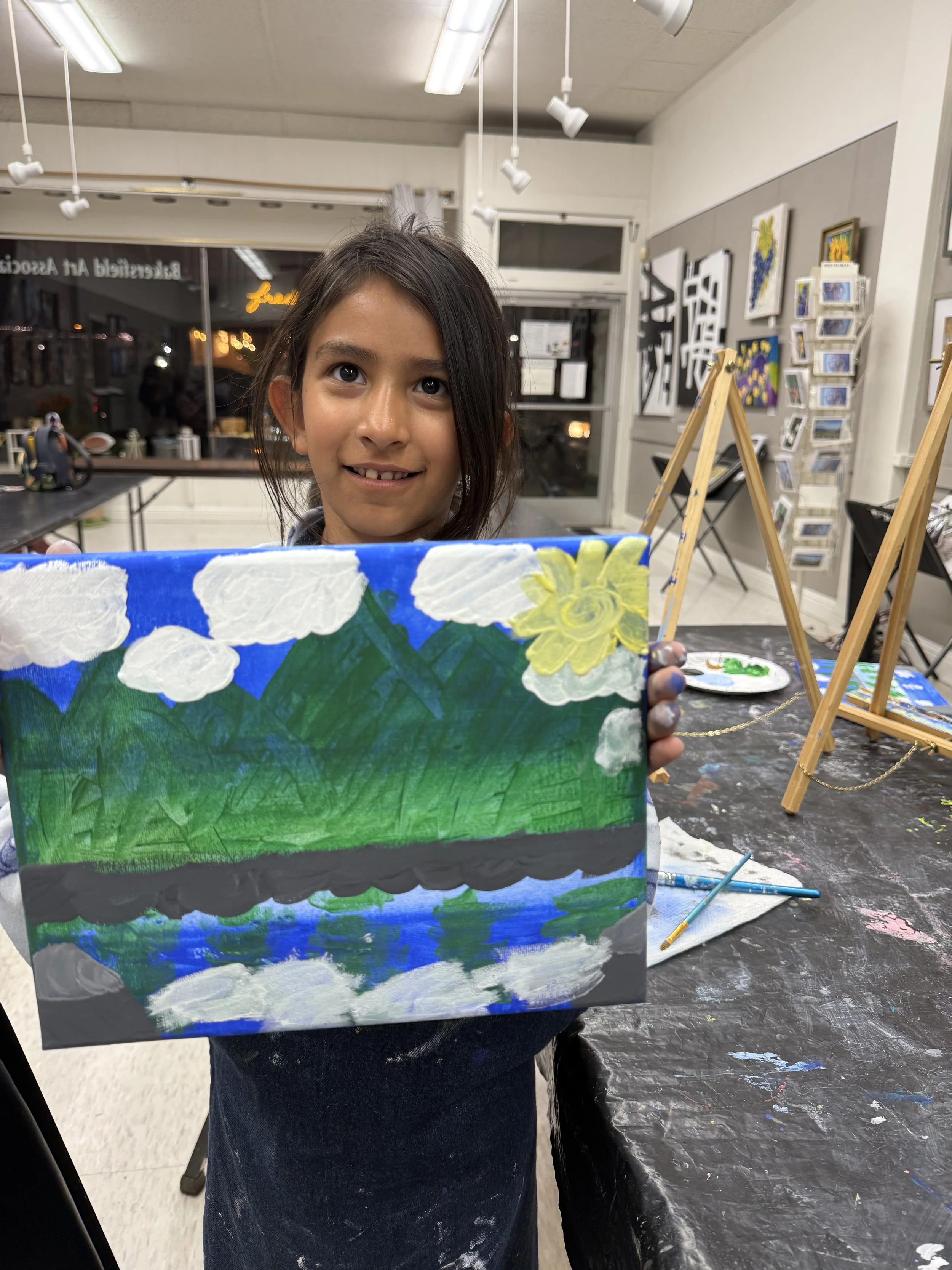 A young girl with dark hair holding a painting of a landscape featuring mountains, a lake, sky, clouds, and a sun, inside an art studio.