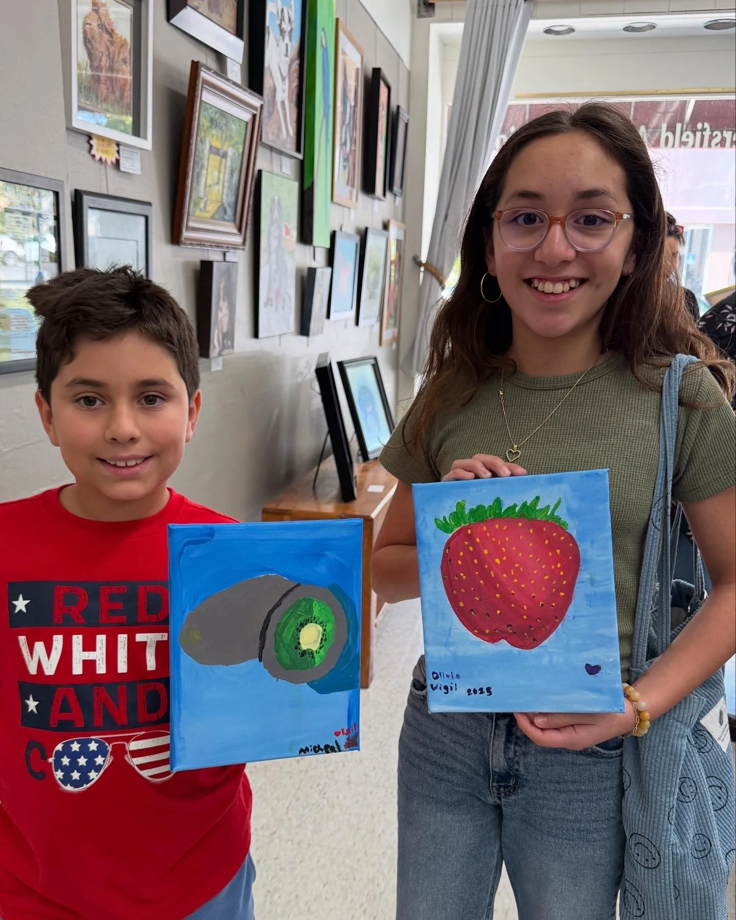 A boy and girl standing indoors, each holding a small canvas painting. The boy's painting depicts a sliced kiwi fruit, and the girl's painting shows a large strawberry. They are smiling and appear to be at an art exhibit or gallery.