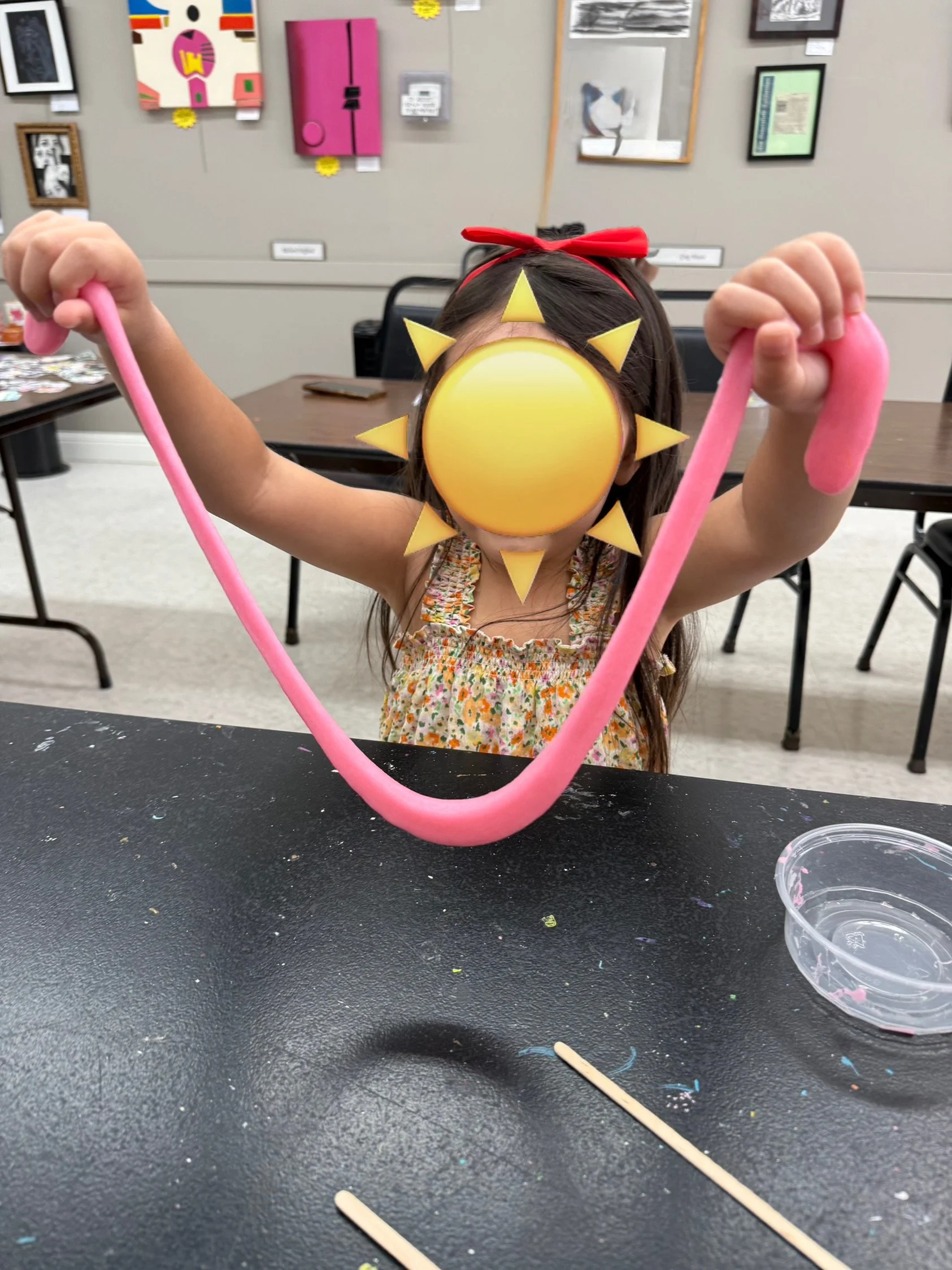 Child with a red headband and floral dress holding up pink slime with a sun emoji overlaying her face, in a classroom or art room setting.